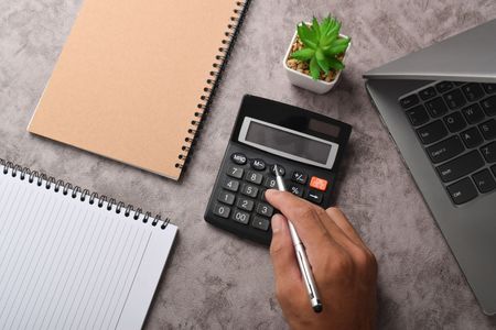 Hand using calculator on a desk with a laptop, notebooks, and a plant.