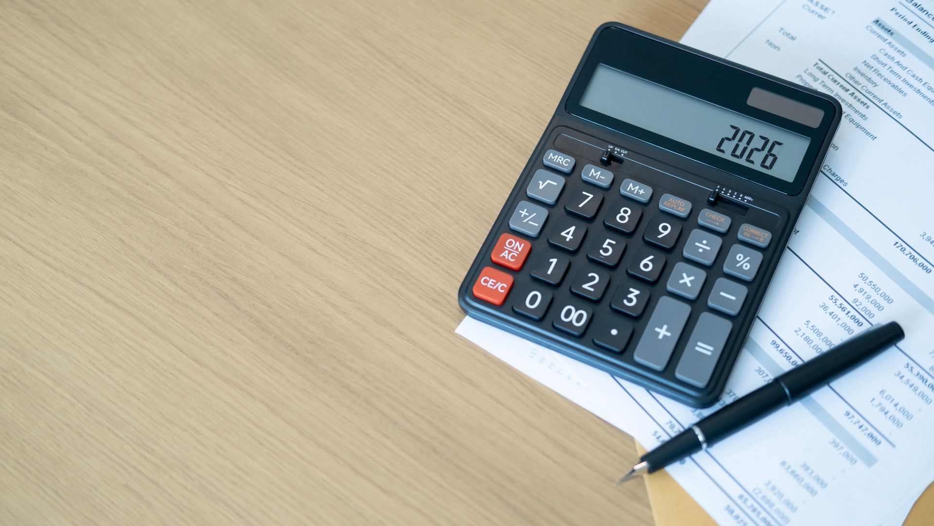 Calculator with 2026 displayed, resting on papers with a pen, on a light wood surface.