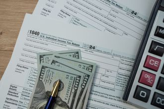 Tax forms, US currency, calculator, and pen on a wooden surface.