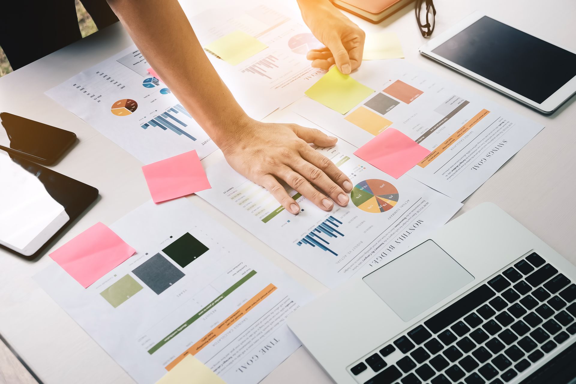 Person reviewing charts with sticky notes, laptop, and tablet on a desk.