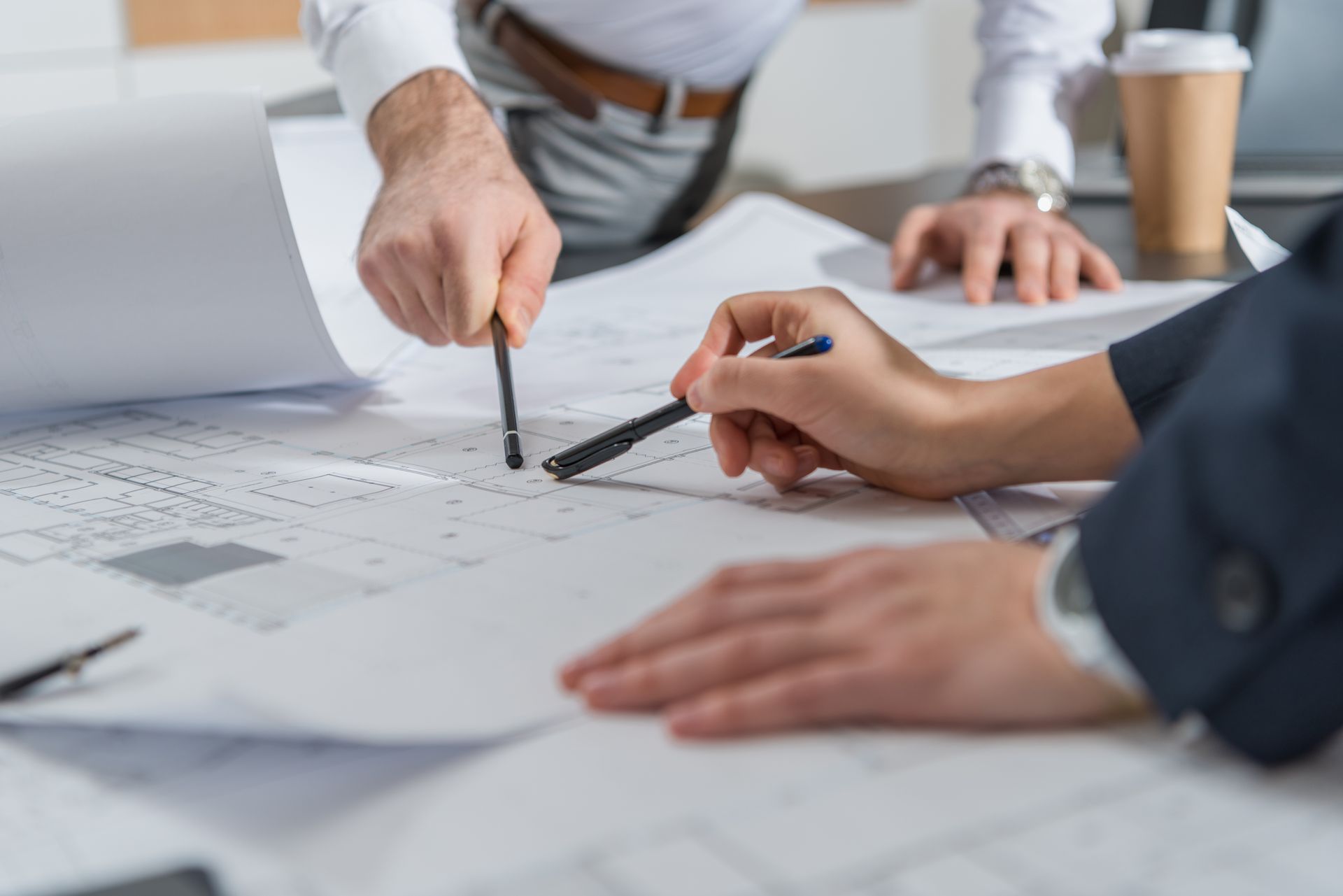 Hands pointing at architectural plans on a table with a laptop and coffee cup nearby