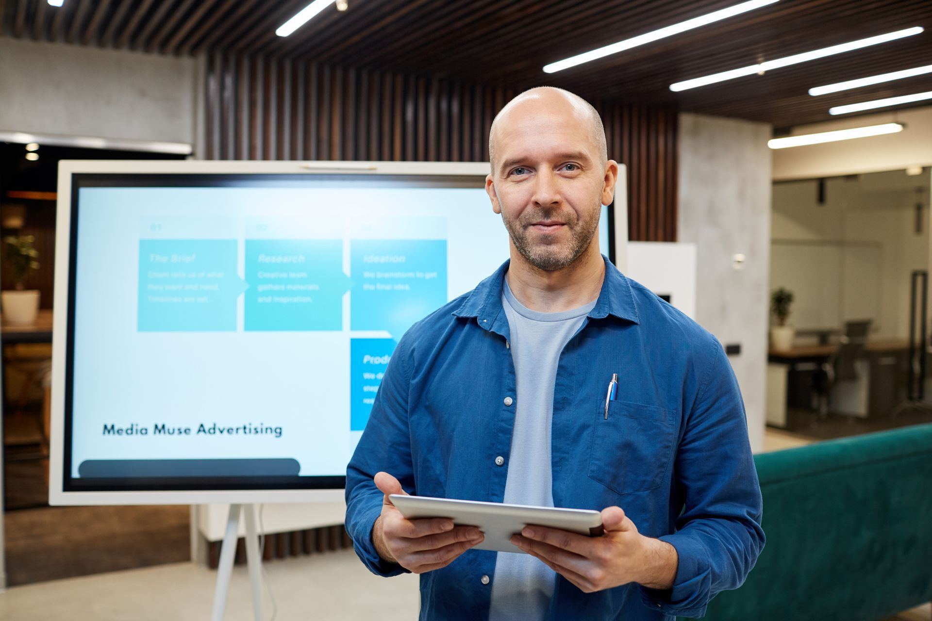 A person in a blue shirt holds a tablet in an office in front of a screen displaying a flowchart about media advertising. A person in a blue shirt holds a tablet in an office in front of a screen displaying a flowchart about media advertising.