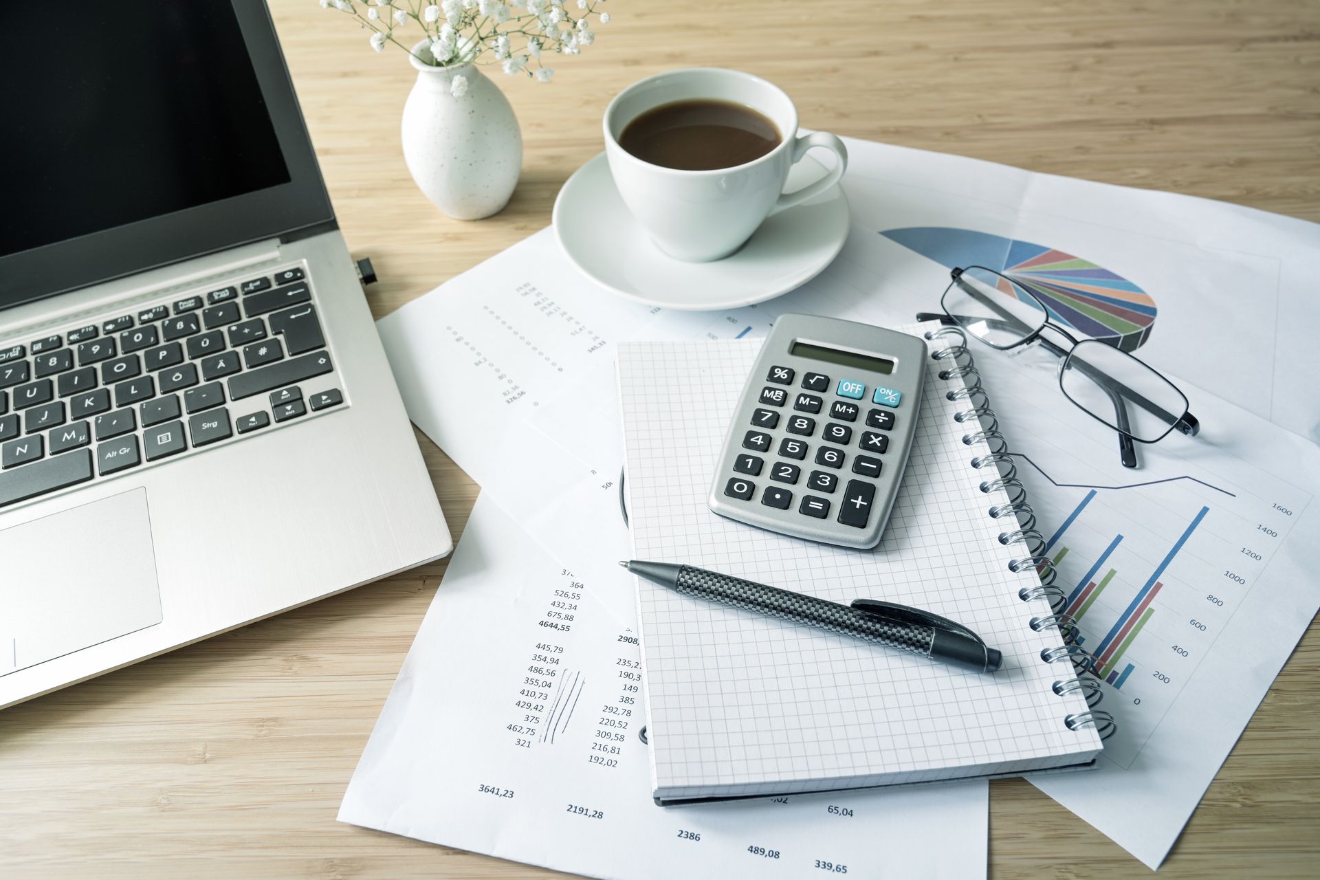 Laptop, coffee cup, calculator, and papers on a wooden desk, possibly for financial analysis.