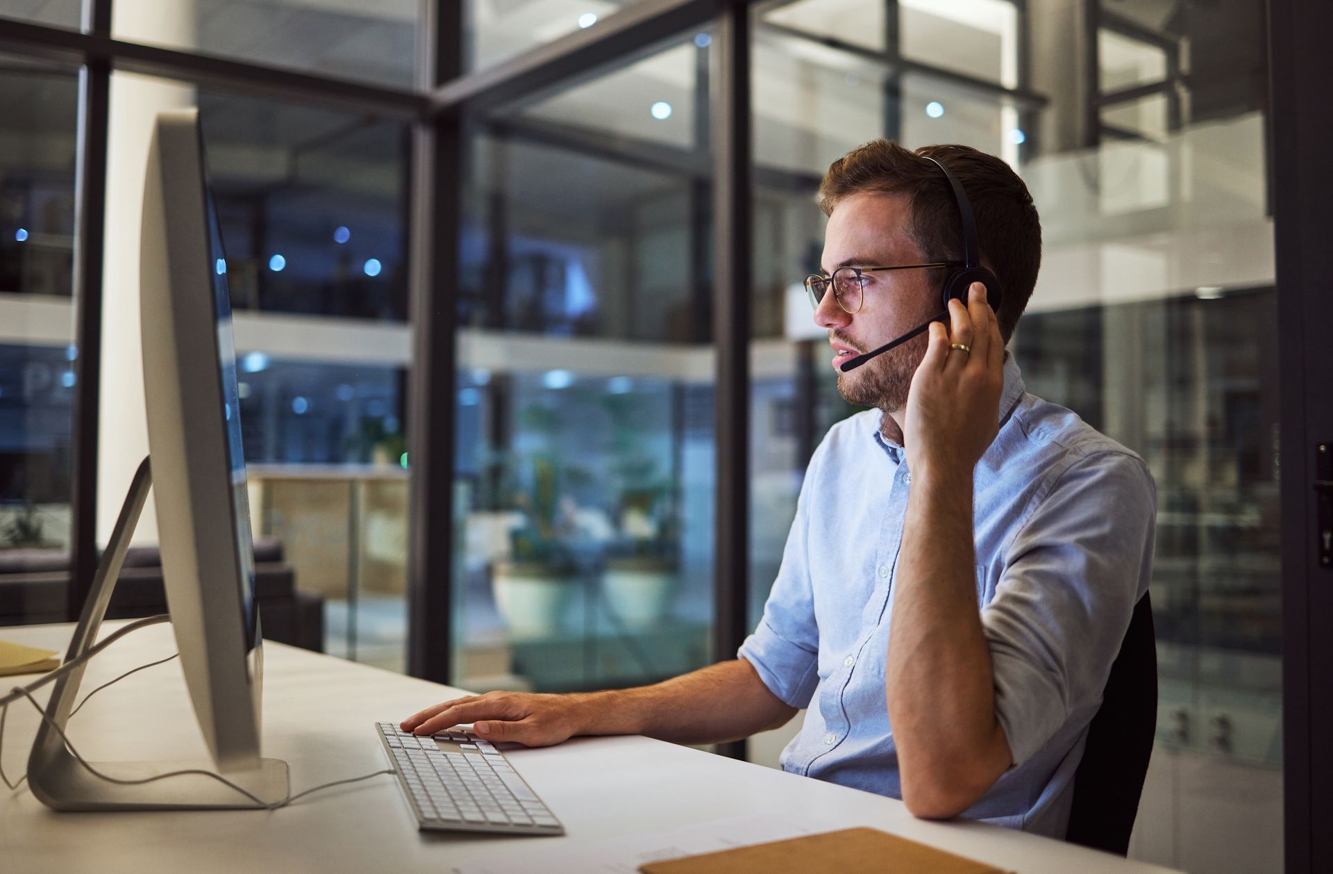 A person wearing a headset works at a computer in a modern office with glass walls, their hand resting on a keyboard. A person wearing a headset works at a computer in a modern office with glass walls, their hand resting on a keyboard.
