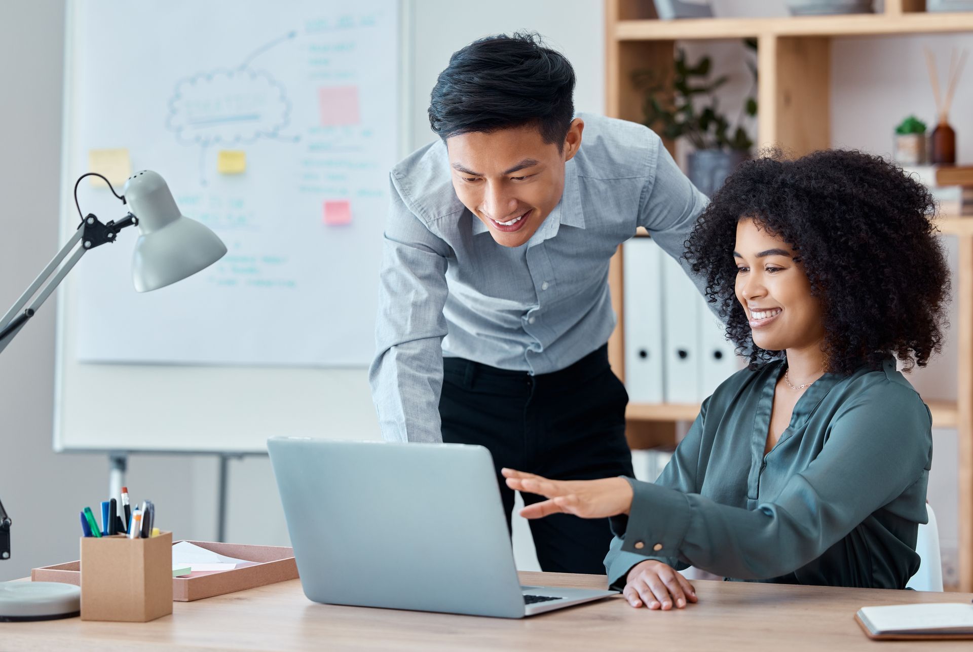 Two colleagues sit at a desk, smiling and collaborating while reviewing information on a laptop in an office setting. Two colleagues sit at a desk, smiling and collaborating while reviewing information on a laptop in an office setting.