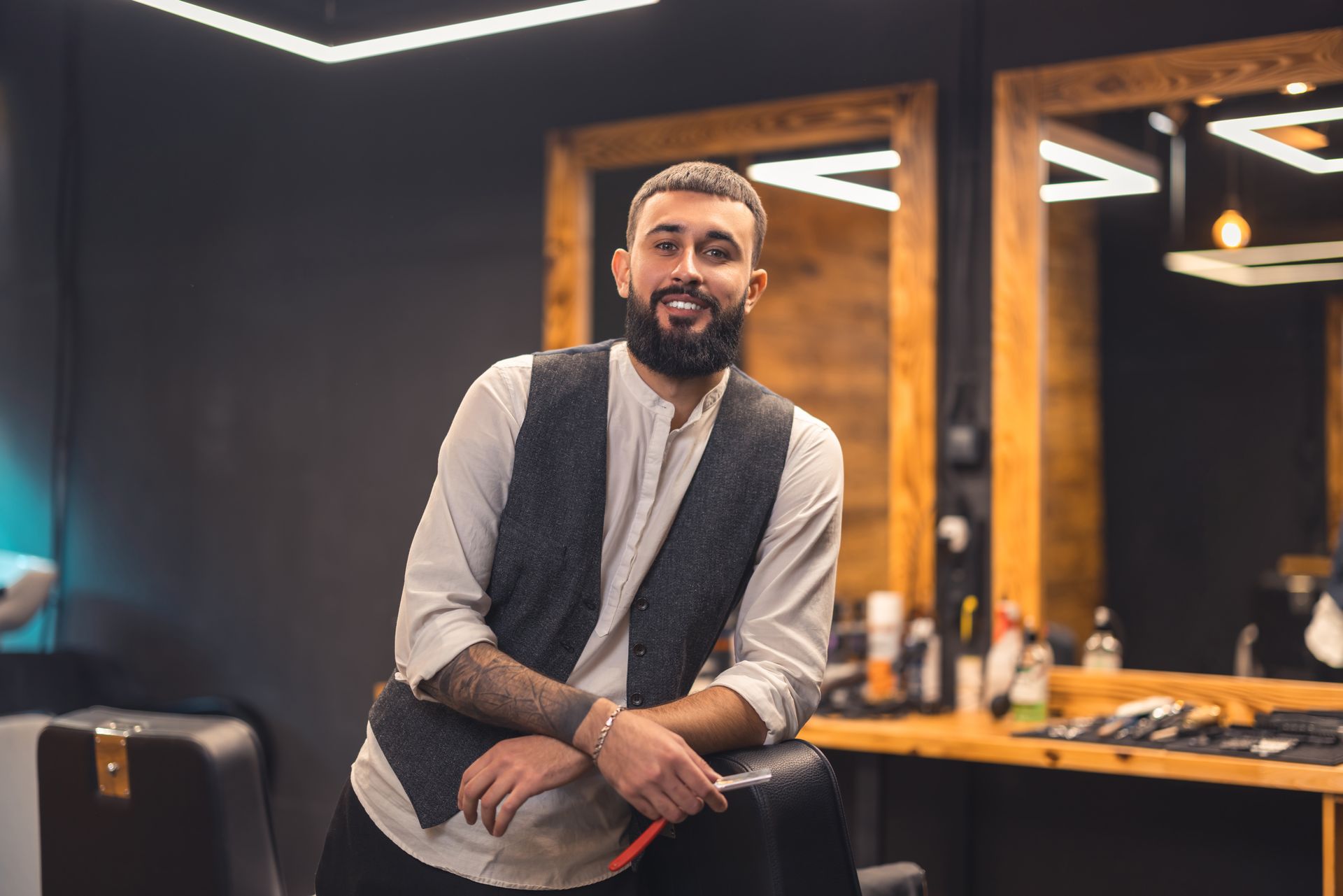 Person using a calculator and pen, writing on paper at a desk. A phone, keyboard, and notebook are also present. A smiling, bearded barber wearing a white shirt and dark vest leans against a salon chair in a wood-accented shop.