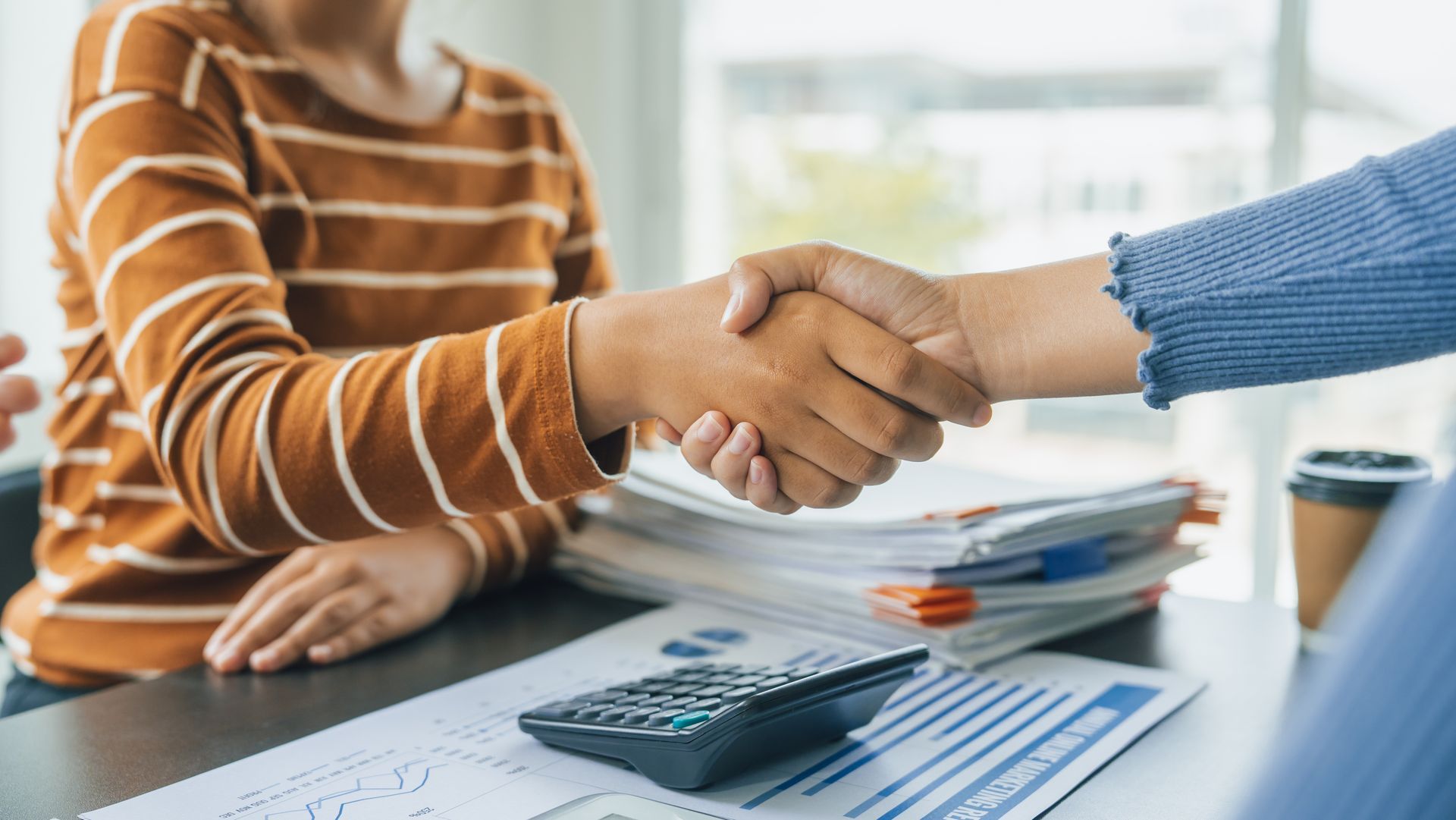 Two people shake hands over a desk with a calculator, documents, and a coffee cup, representing a business agreement.