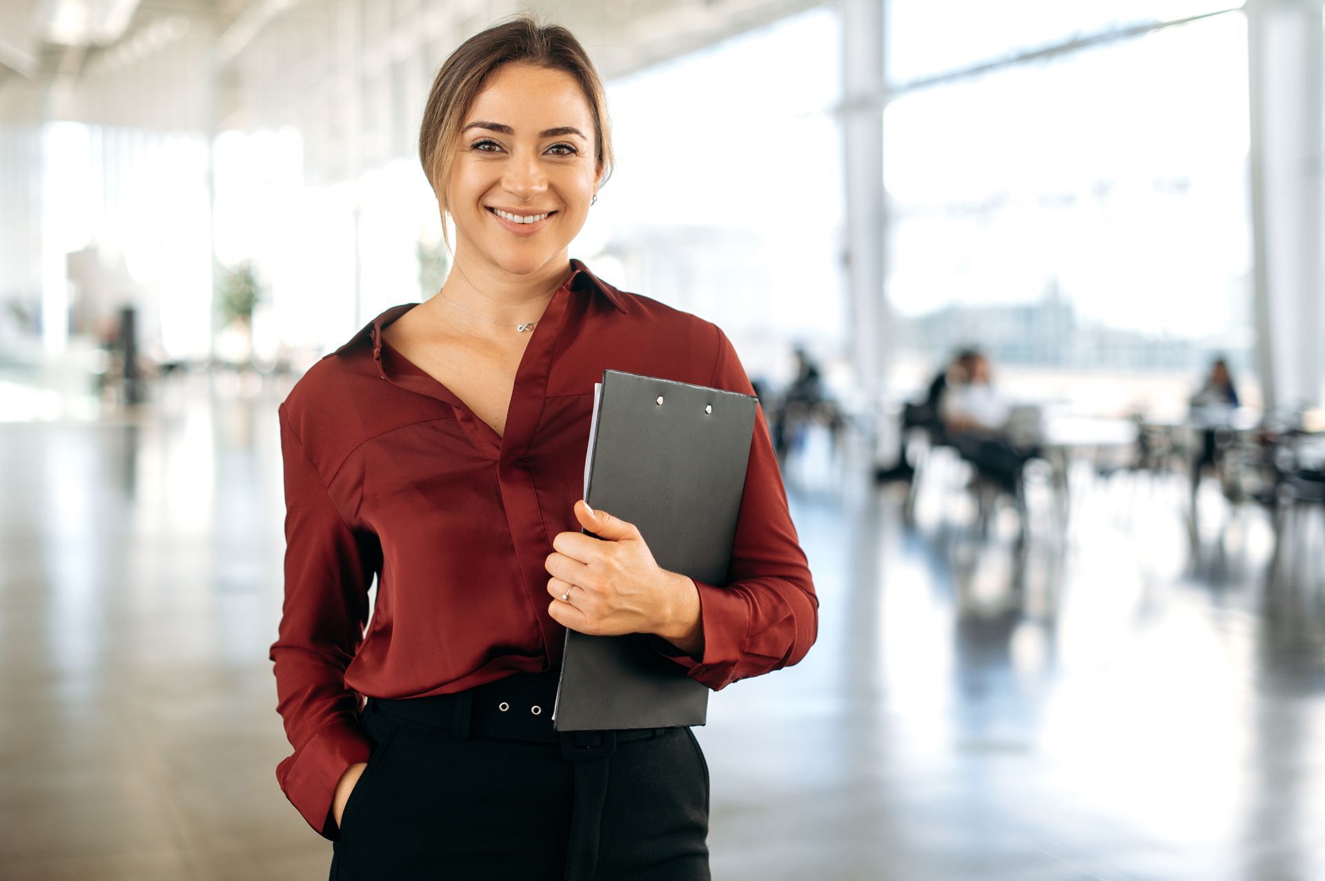 A person in a maroon blouse and dark pants stands in a bright office, smiling and holding a black clipboard. A person in a maroon blouse and dark pants stands in a bright office, smiling and holding a black clipboard.