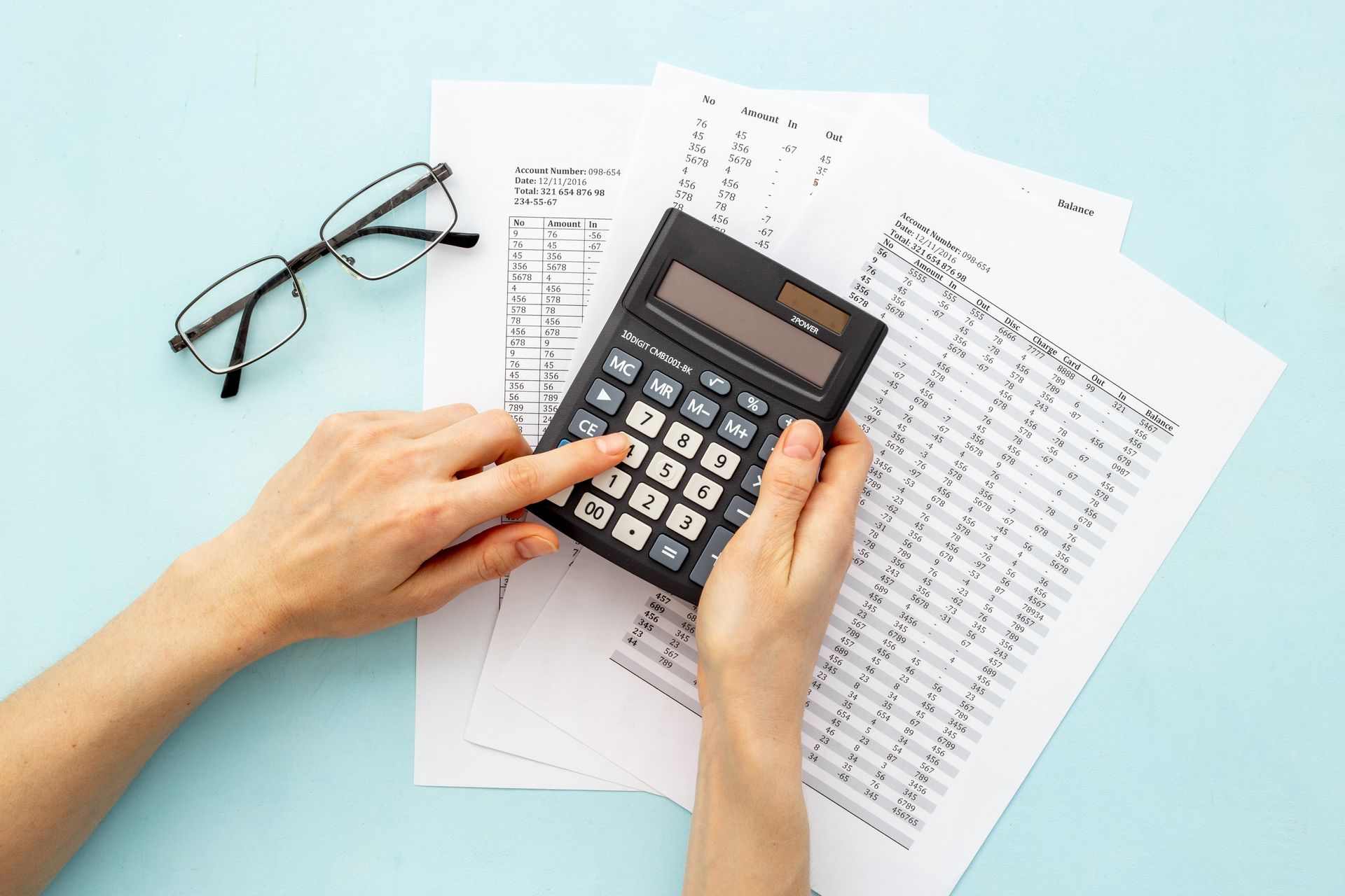Hands using calculator, papers and eyeglasses on a blue background.