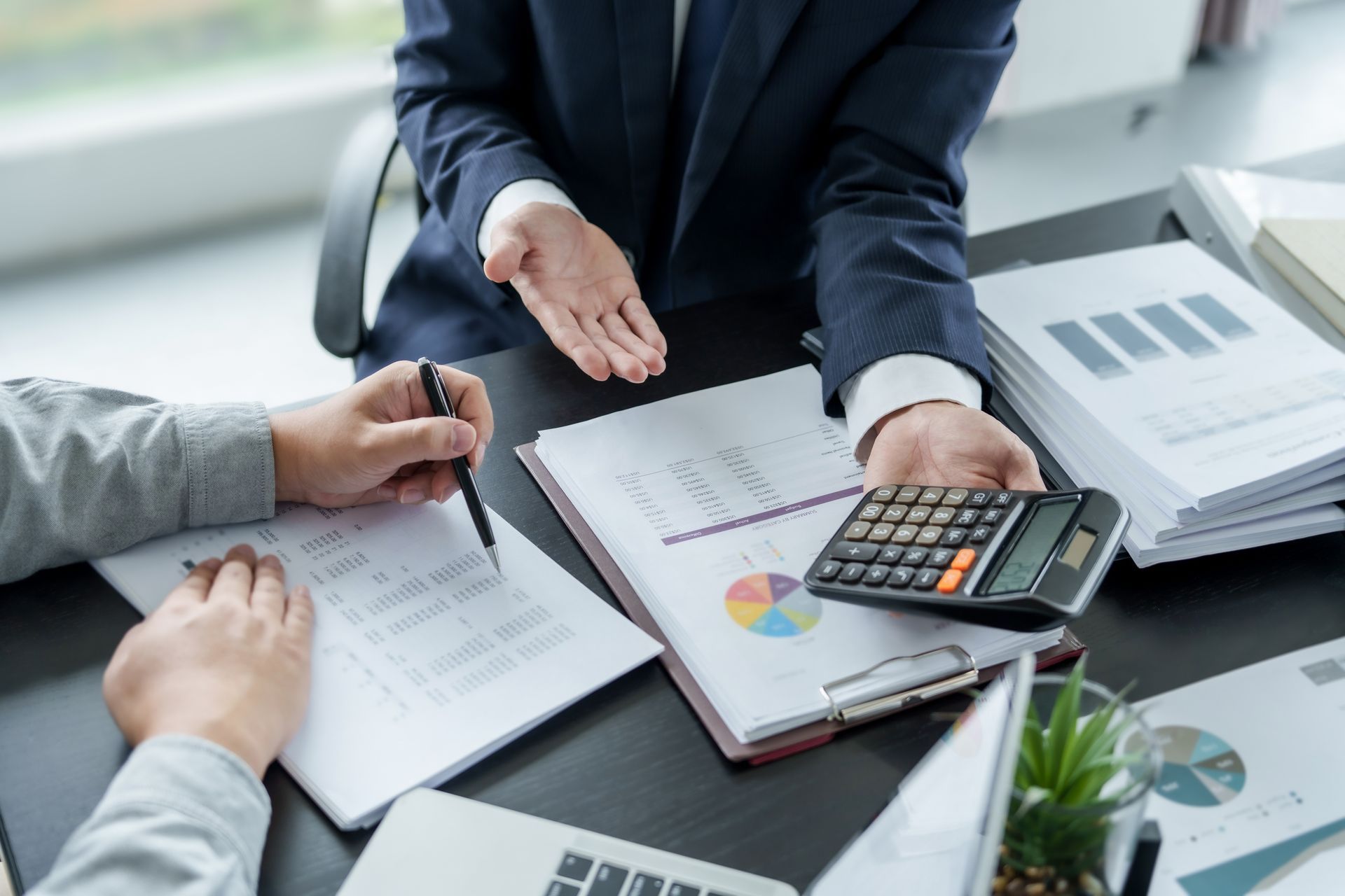 Two people reviewing financial documents, one using a calculator, in an office setting. Two people reviewing financial documents, one using a calculator, in an office setting.