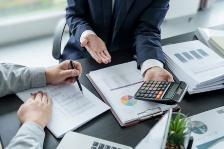 Two people reviewing financial documents, one using a calculator, in an office setting.