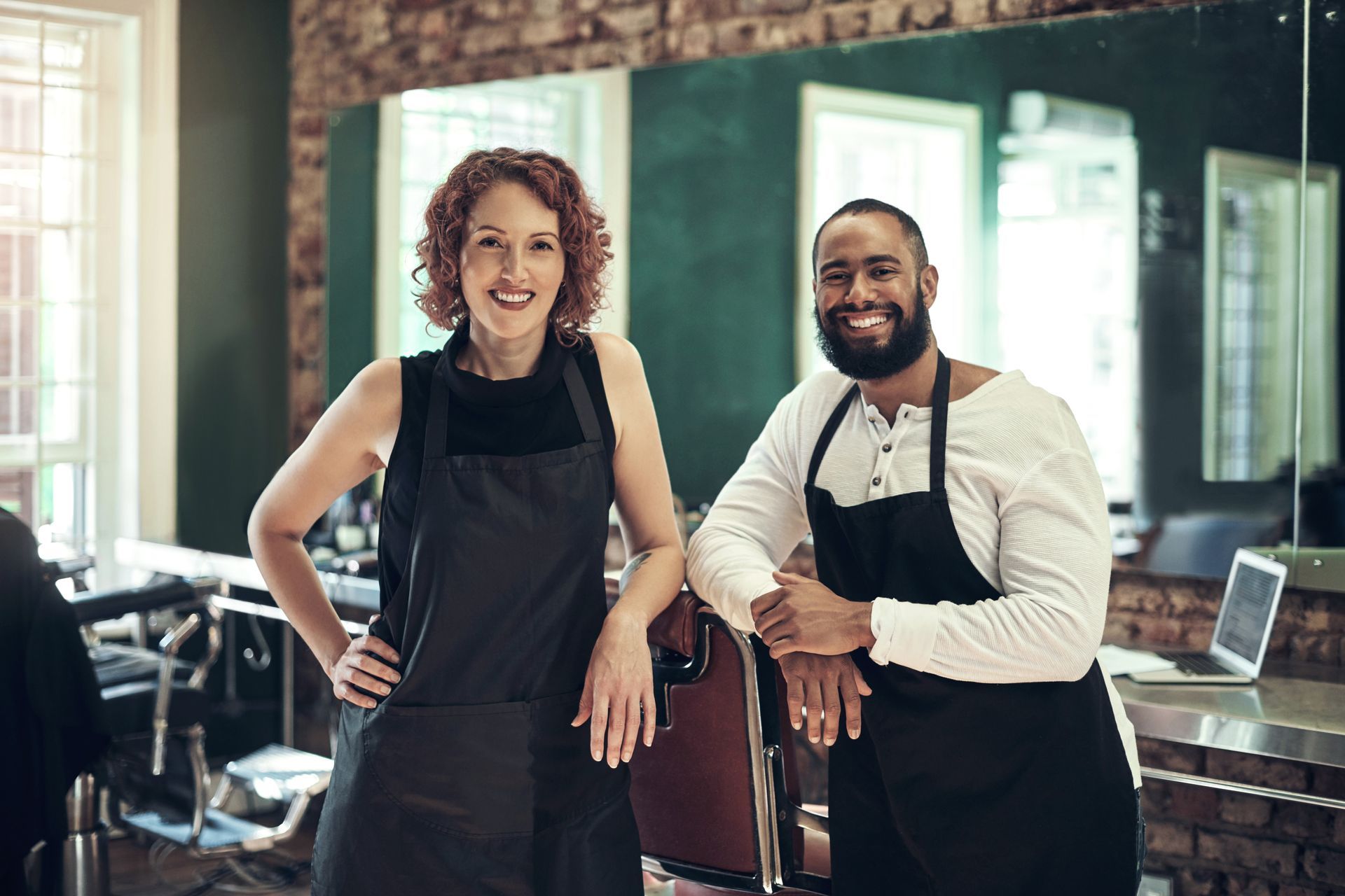 Person points at receipts with calculator, budgeting expenses on a white desk. Two smiling salon staff wearing black aprons stand in a studio with brick walls and barber chairs.