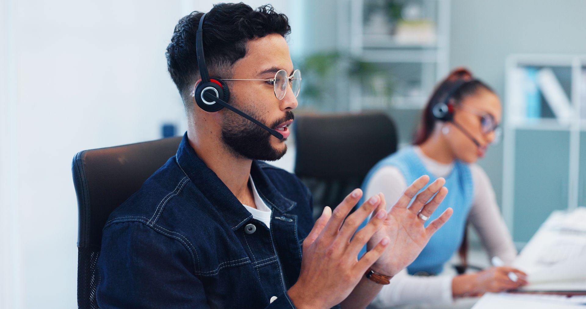 A team member in a headset speaks with hand gestures in a call center office while a colleague works in the background. A team member in a headset speaks with hand gestures in a call center office while a colleague works in the background.