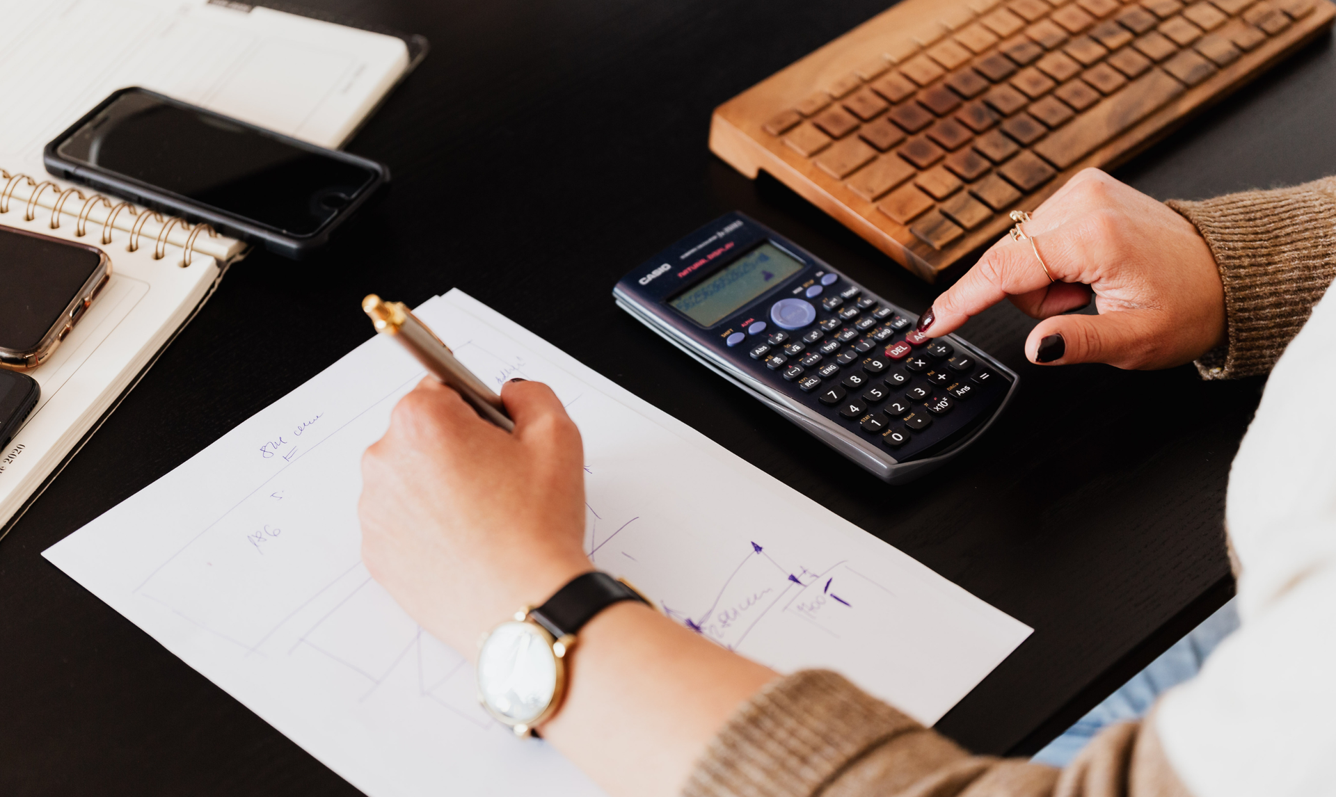 Person using a calculator and pen, writing on paper at a desk. A phone, keyboard, and notebook are also present.