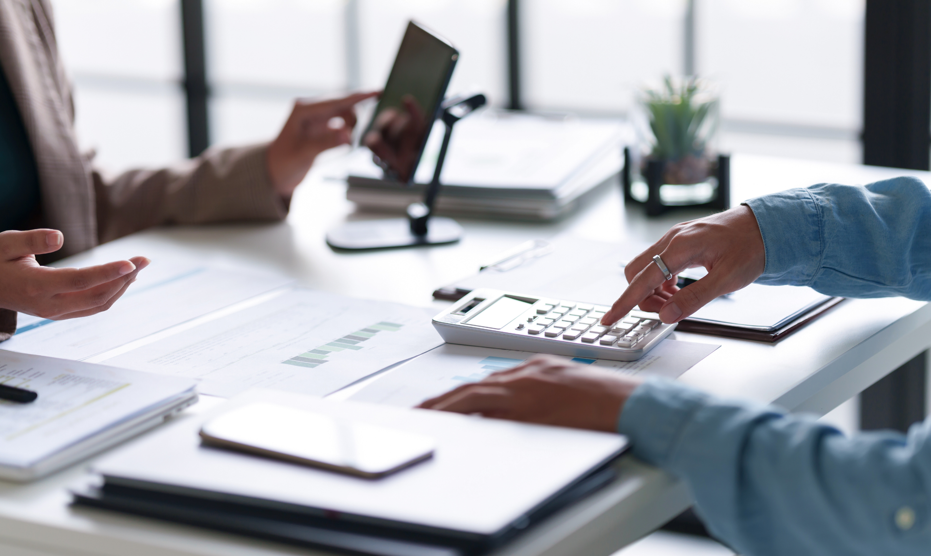 Two people at a desk, reviewing documents and using a calculator. A tablet and a smartphone are also present.