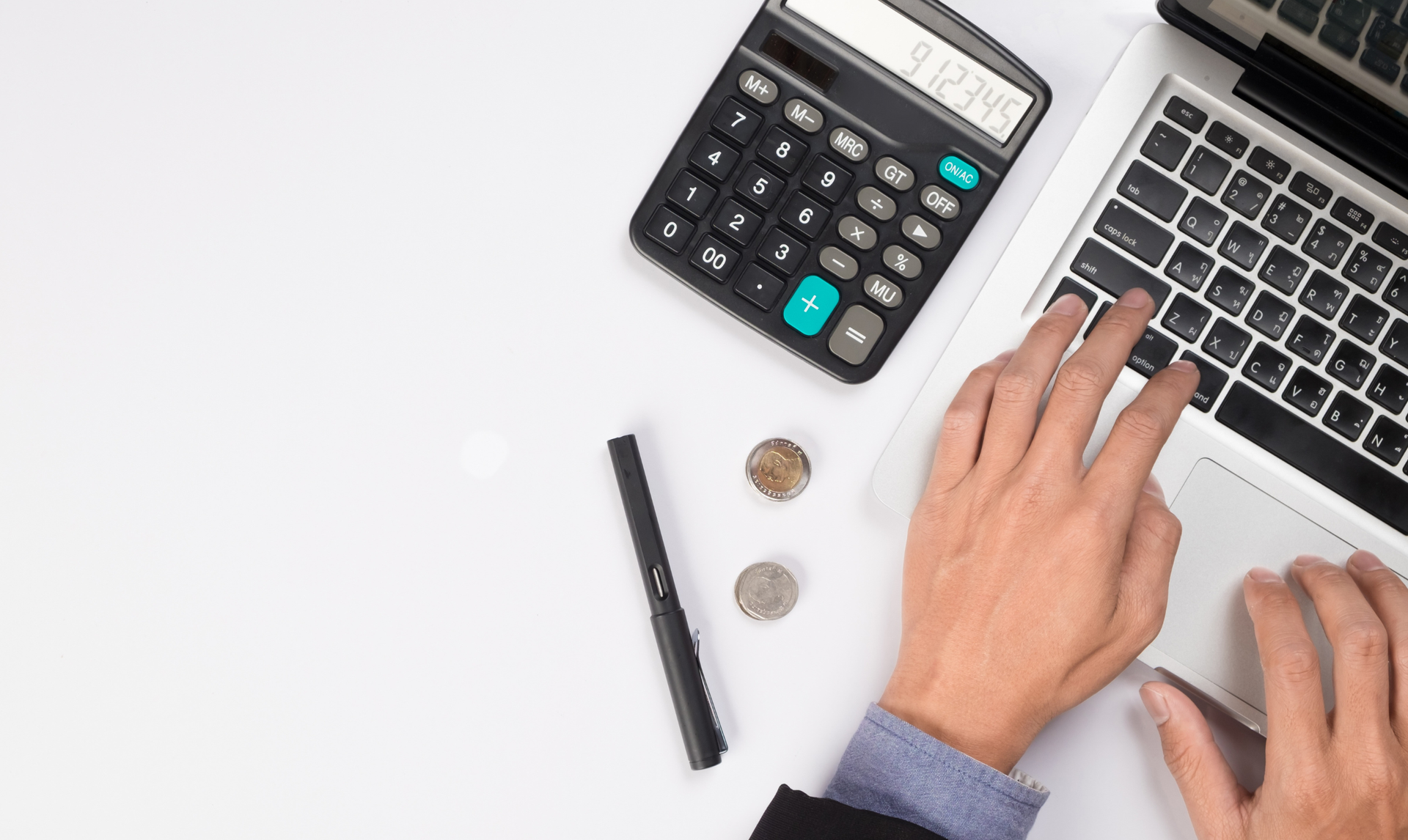 Hands typing on a laptop with a calculator, pen, and coins on a white surface.