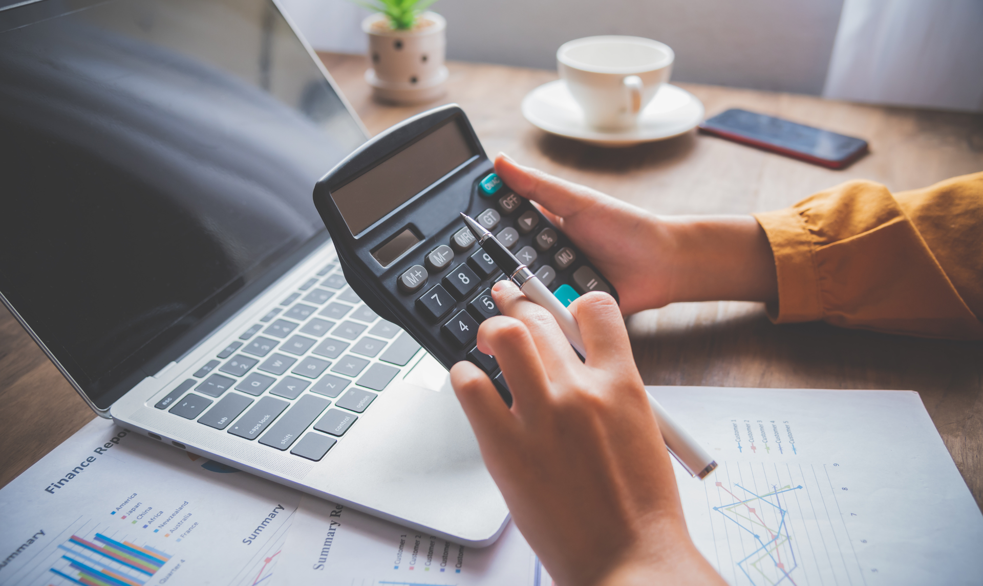 Person using a calculator with a laptop, documents, and coffee cup nearby.