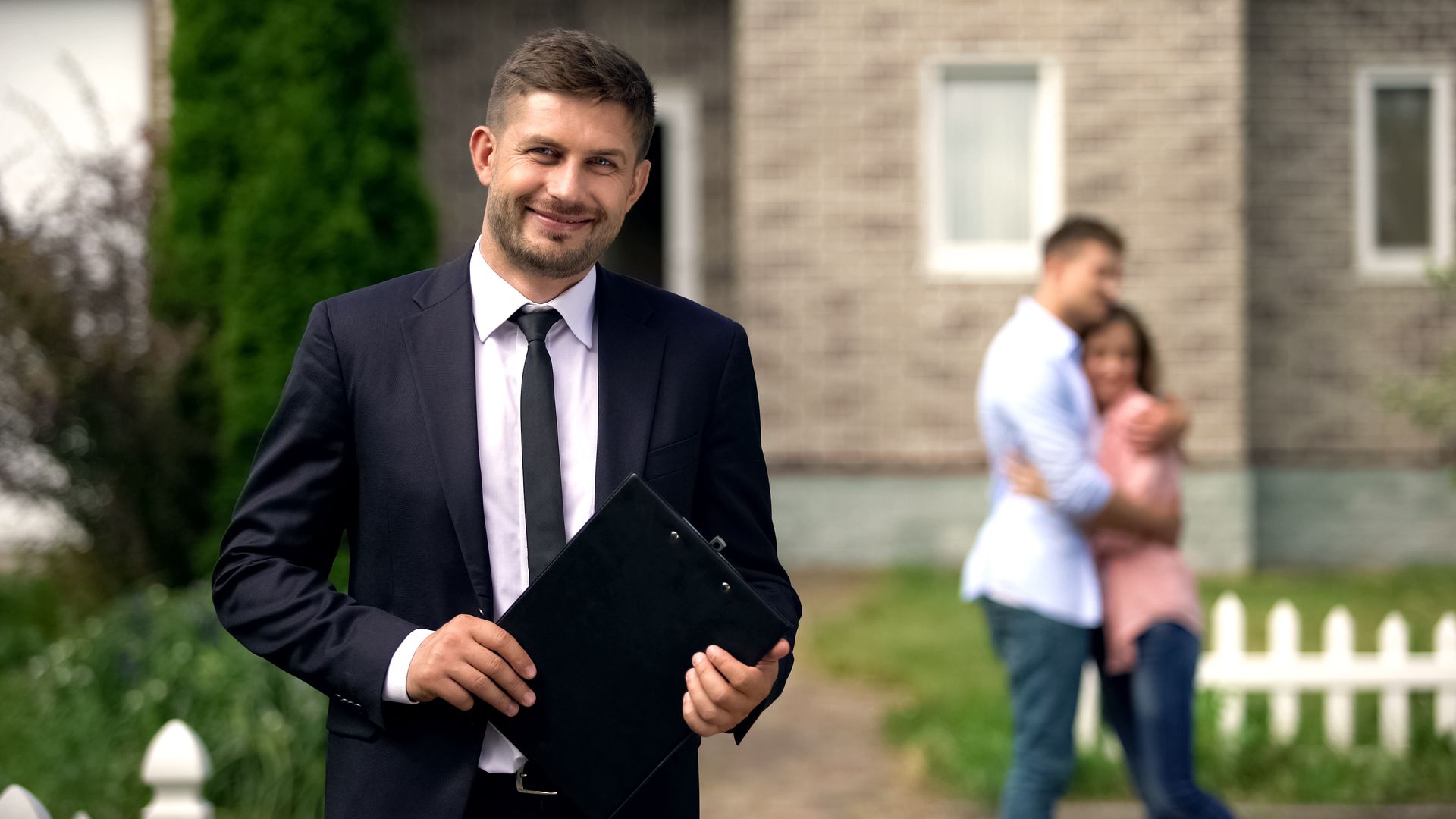 A real estate agent holding a clipboard in front of a home with clients in the background. A real estate agent holding a clipboard in front of a home with clients in the background.