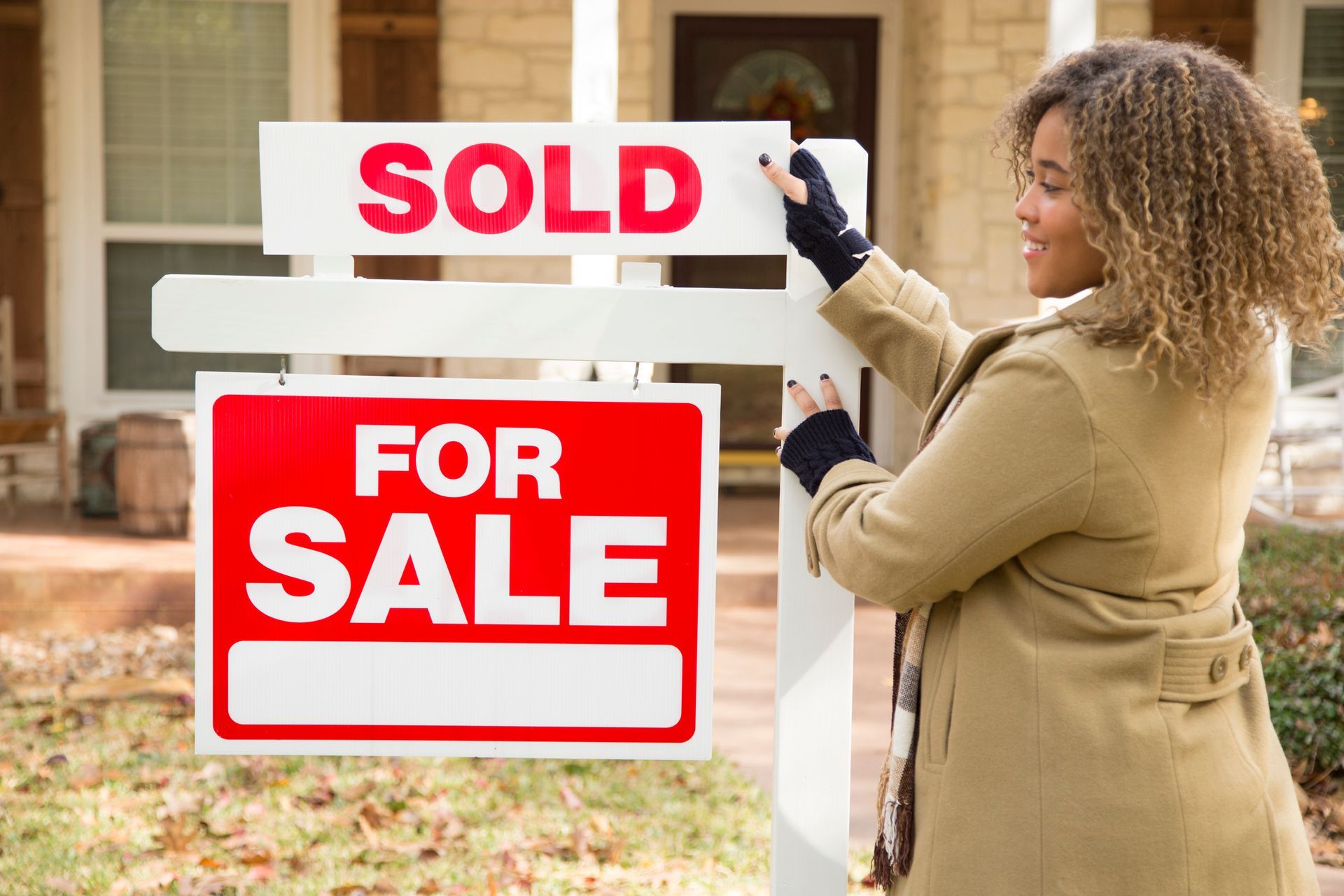 A woman stands in front of a home with a 'Sold' sign, showcasing a local realtor company's services. A woman stands in front of a home with a 'Sold' sign, showcasing a local realtor company's services.