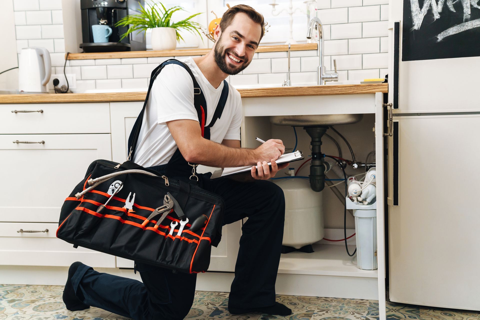 Plumber kneeling by an open sink cabinet, writing on a clipboard with a tool bag nearby.