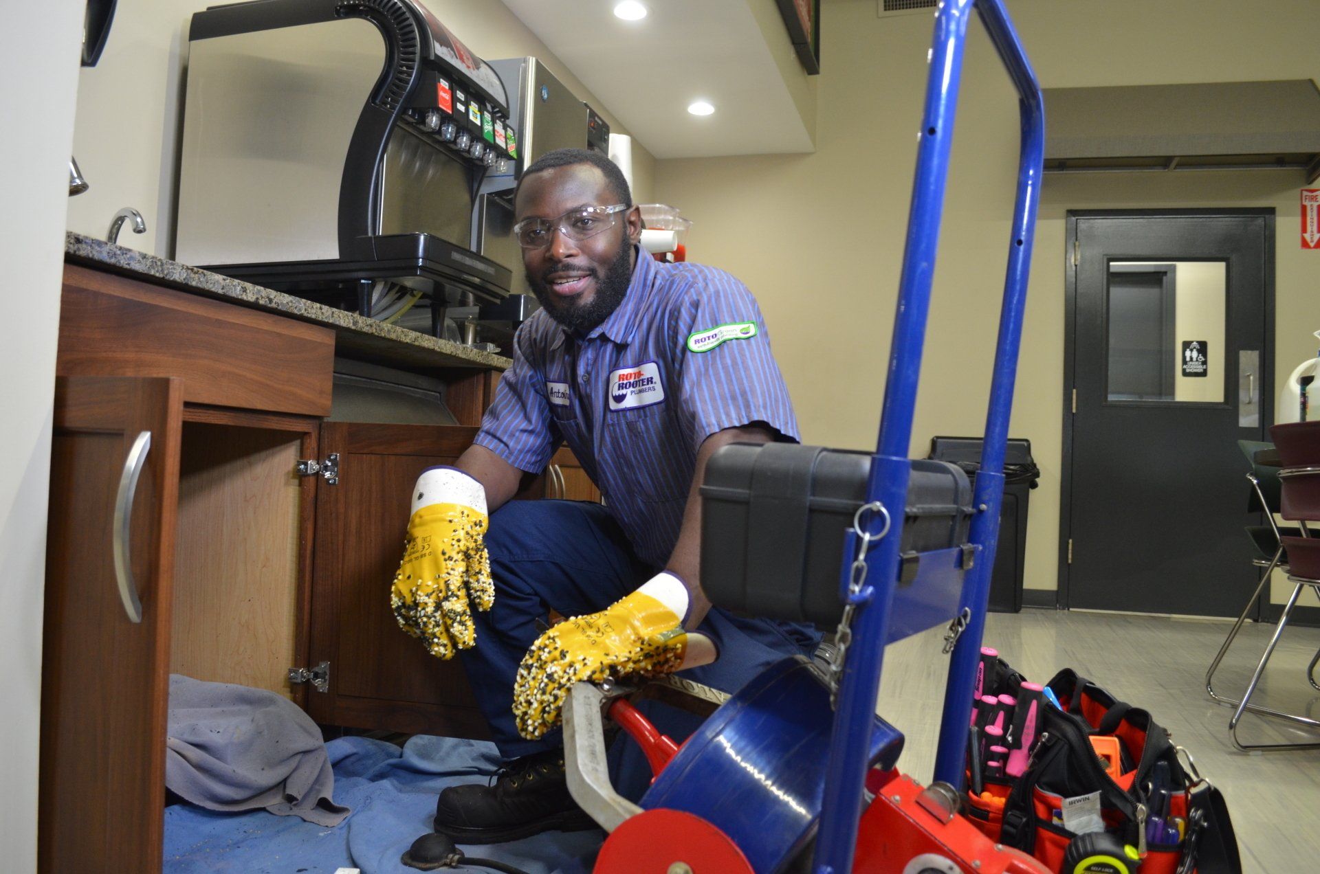 A man in a blue shirt is kneeling down under a sink with a hose in his hand.