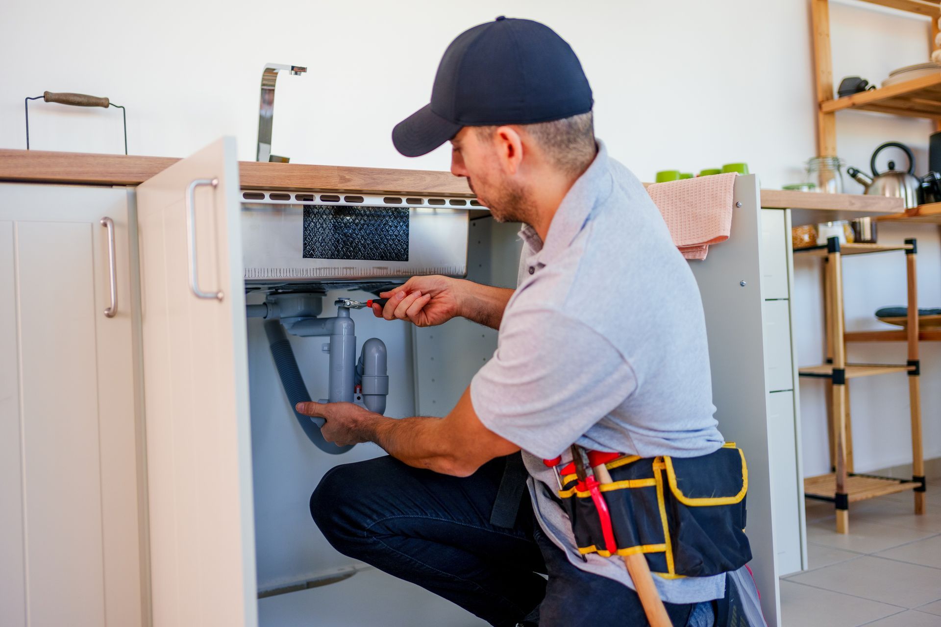 A residential plumbing contractor, wearing a blue hat and gray uniform, works under a kitchen sink.