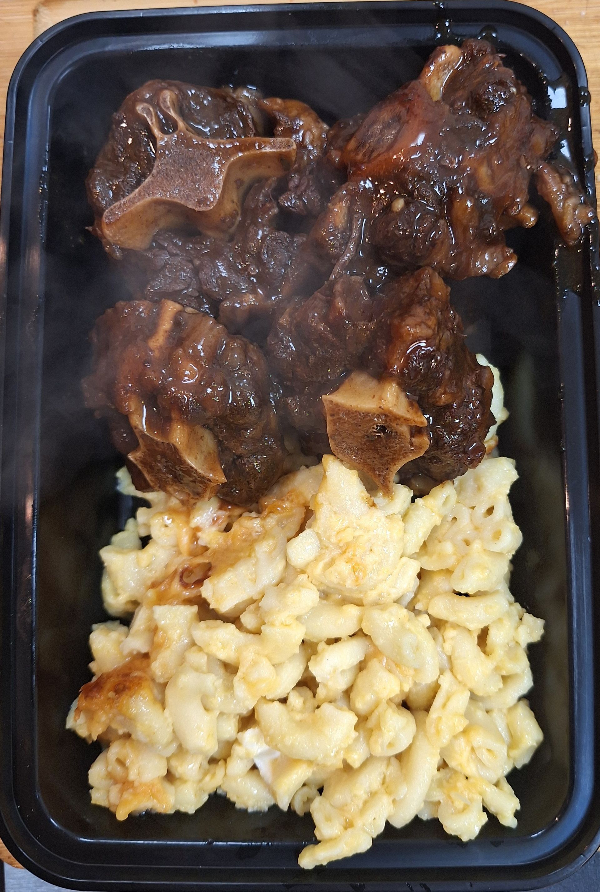 Oxtails with glossy sauce over a portion of egg noodles, served in a black container.