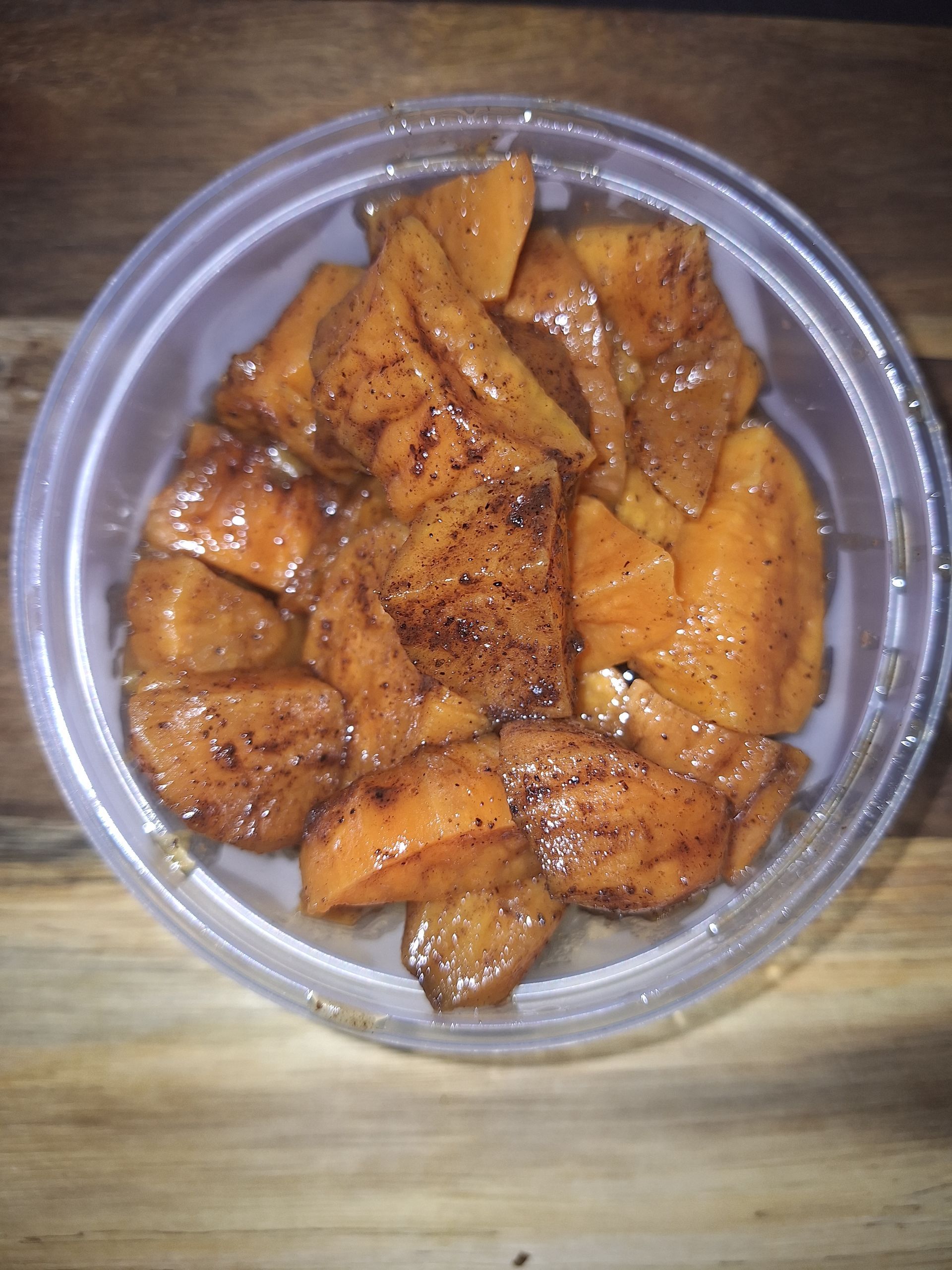 Caramelized sweet potatoes in a clear plastic container, sprinkled with cinnamon, on a wood surface.