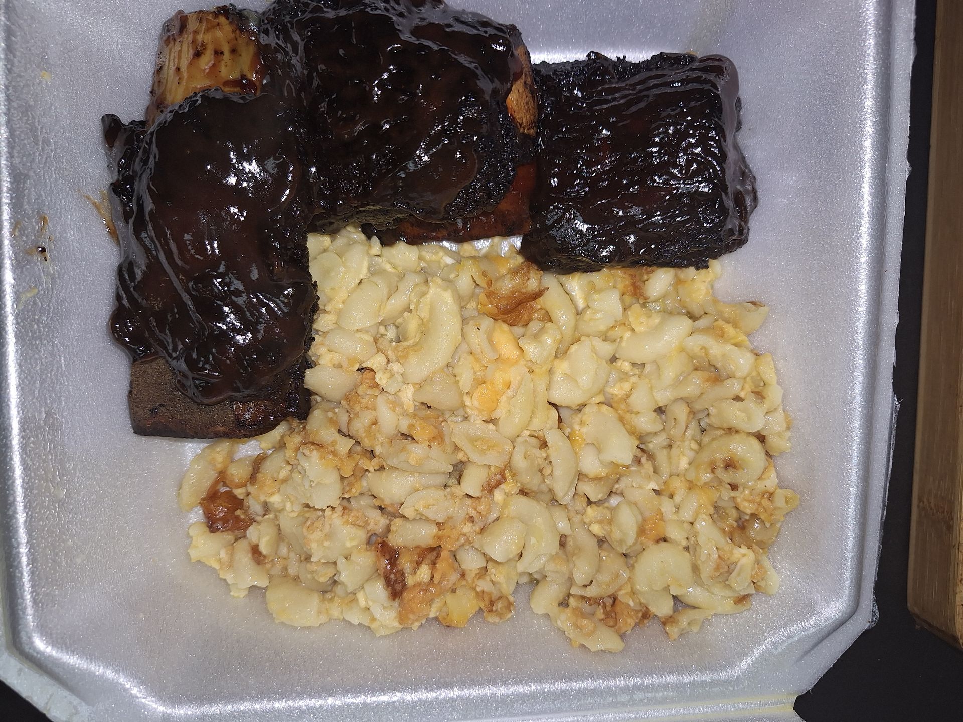 BBQ ribs with a dark, glossy sauce, served with mac and cheese in a white foam container.