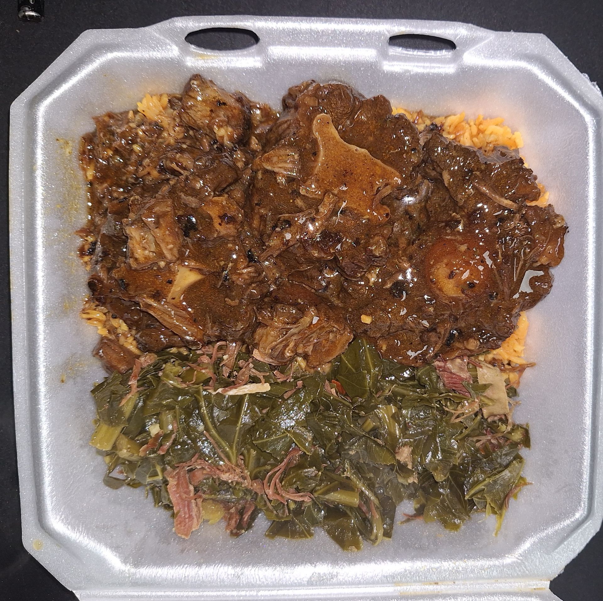 Oxtail dish with rice and collard greens in a white takeout container.