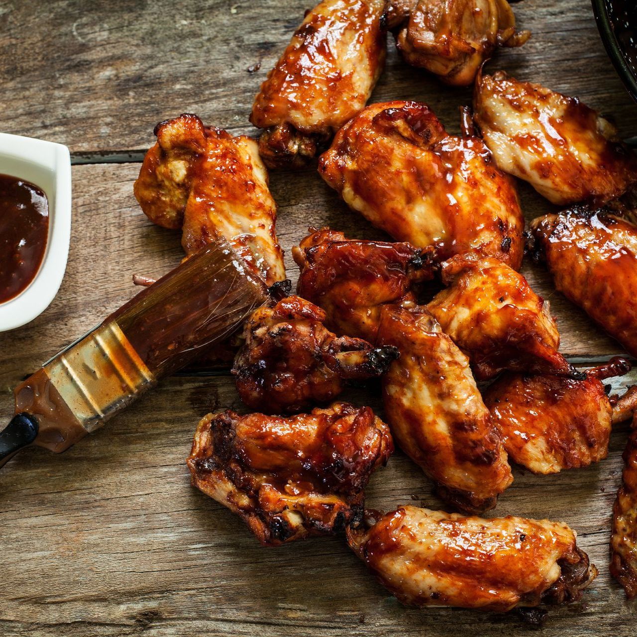 Chicken wings with glaze and brush on wood table, small bowl of sauce.