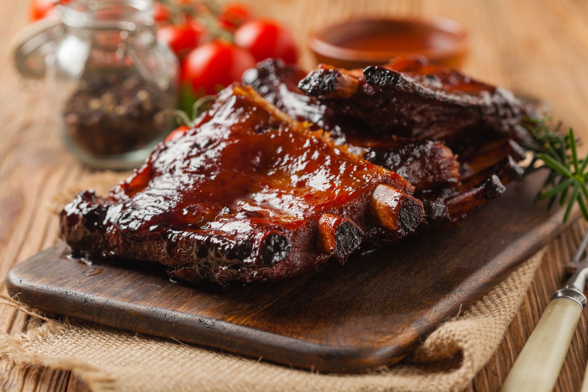 BBQ ribs on a wooden cutting board, glazed in sauce, with tomatoes and spices in the background.