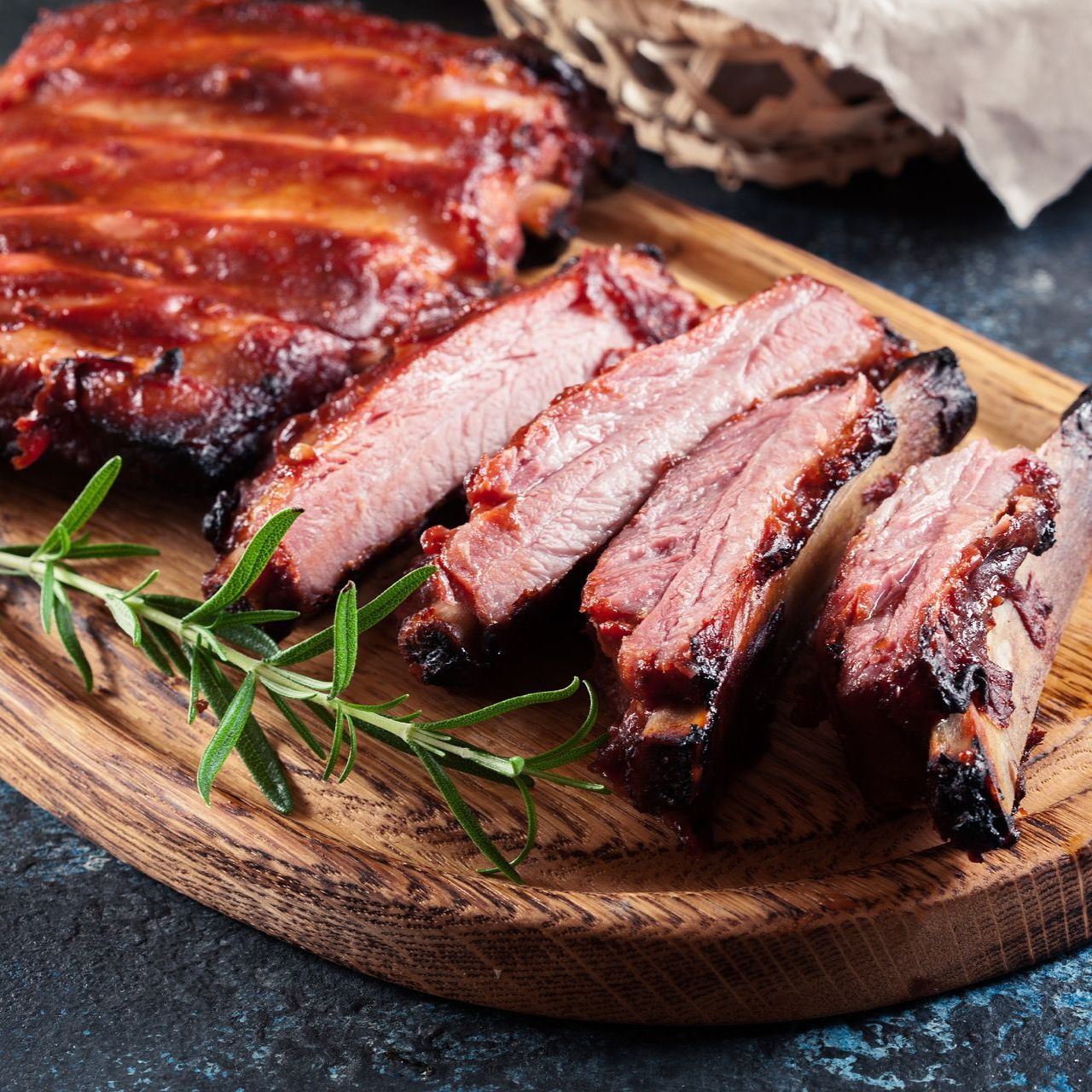 Sliced barbecue ribs on a wooden board with rosemary sprig, dark background.