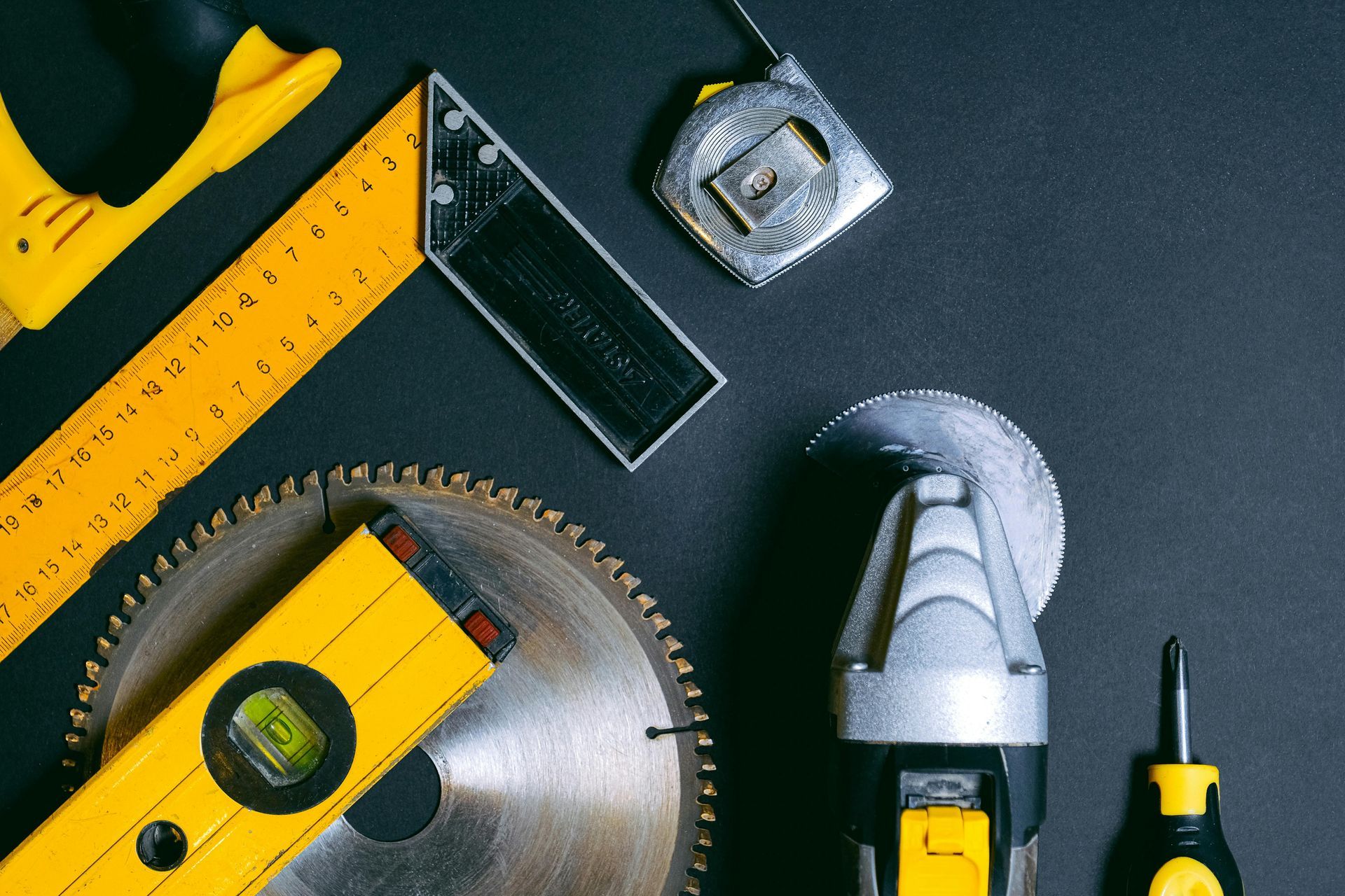 Yellow and silver tools arranged on a black surface, including a saw blade, tape measure, and level.
