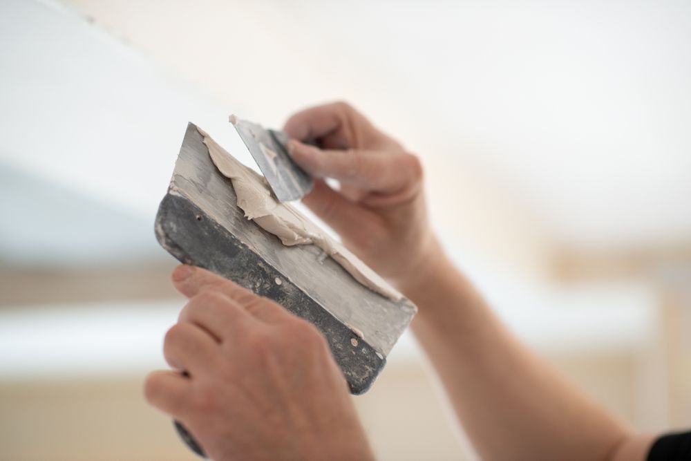 Hands using a putty knife to apply or remove drywall compound on a ceiling.