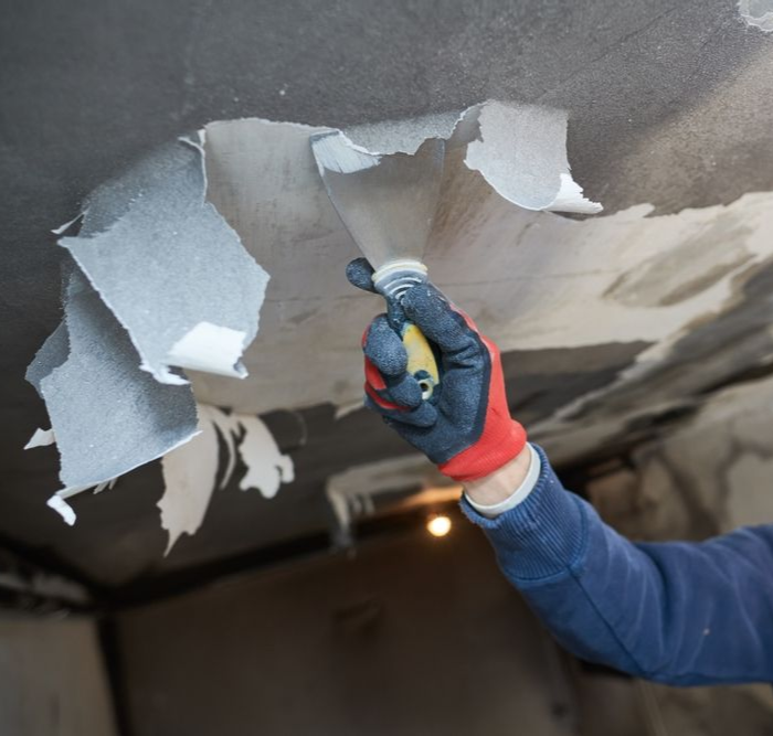Person peeling damaged ceiling material with a scraper, wearing work gloves.