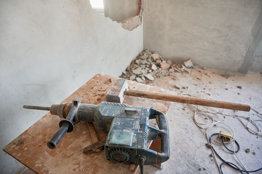 Construction scene with a drill and sledgehammer on a wooden board, debris on the floor and wall.