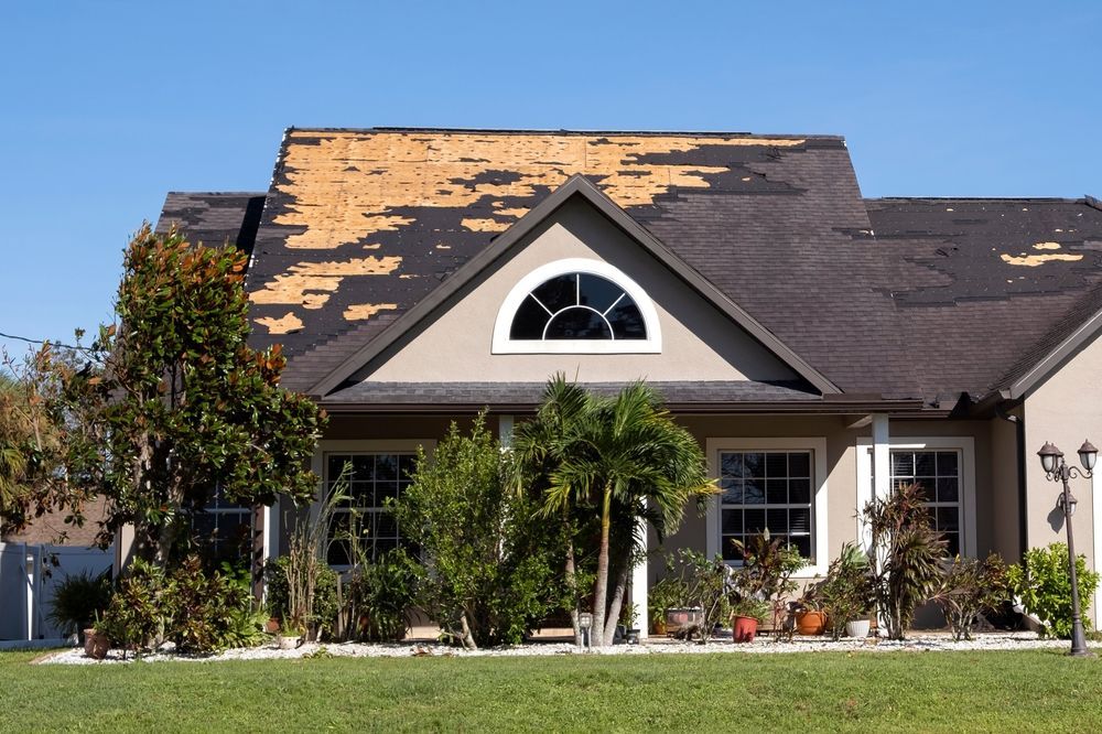 House with significant roof damage, visible peeling shingles against a clear blue sky.
