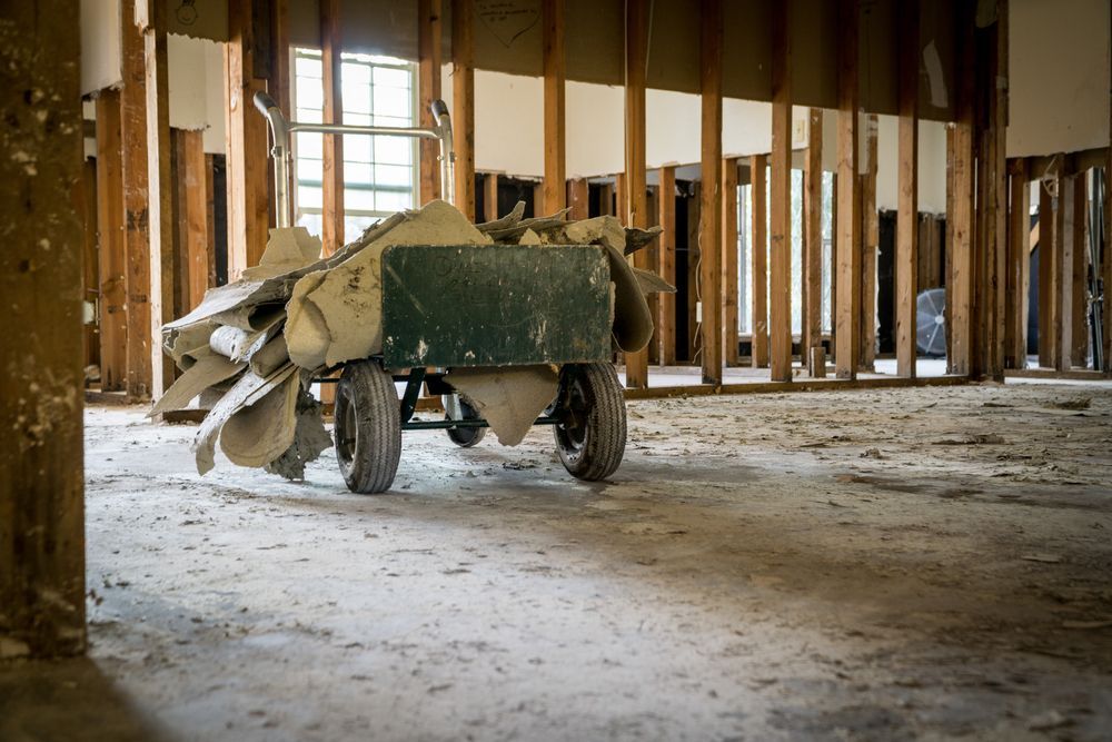 Wheelbarrow filled with debris in a gutted room with exposed wooden framing and a window.