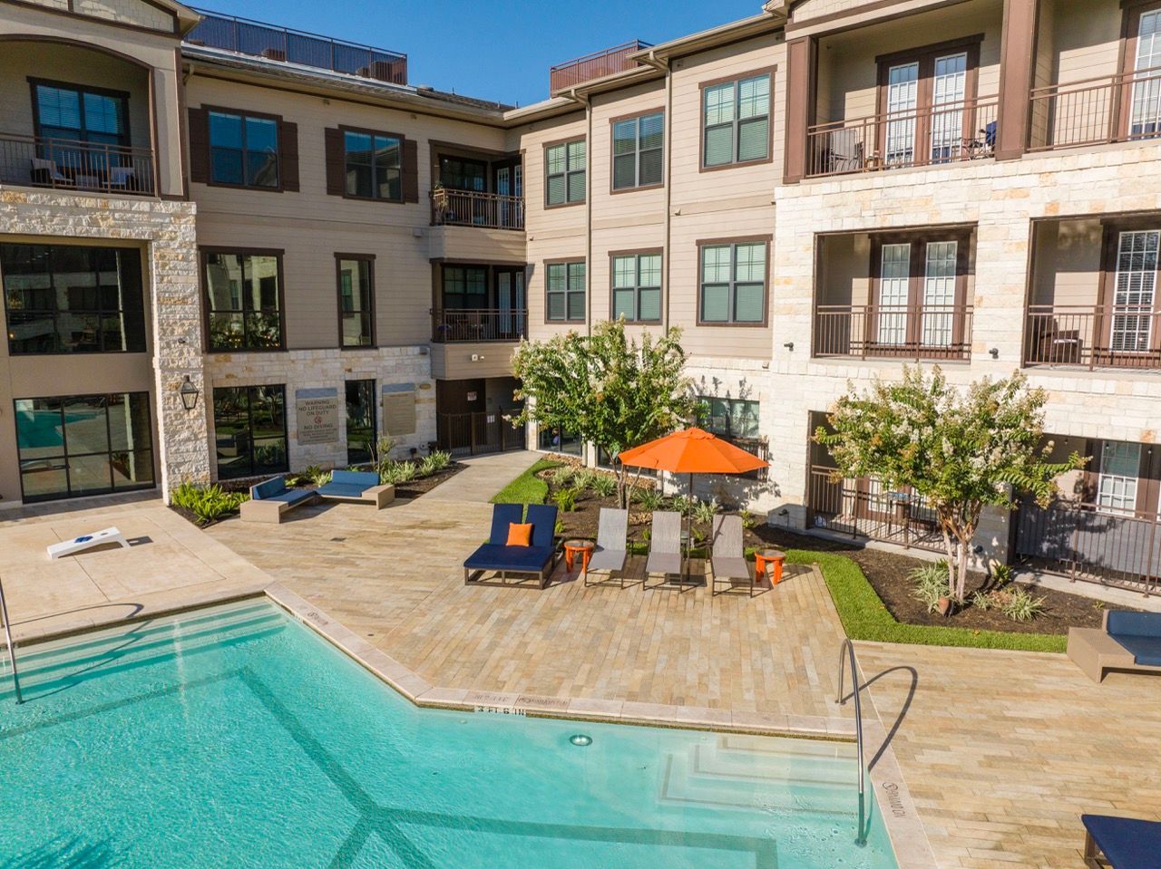 Outdoor pool area with lounge chairs and an orange umbrella in a modern apartment courtyard.