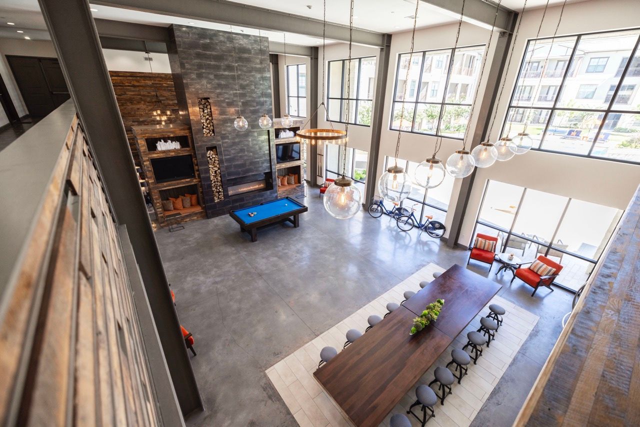 Open-concept community lounge with a long wooden table, stools, fireplace, and large windows.