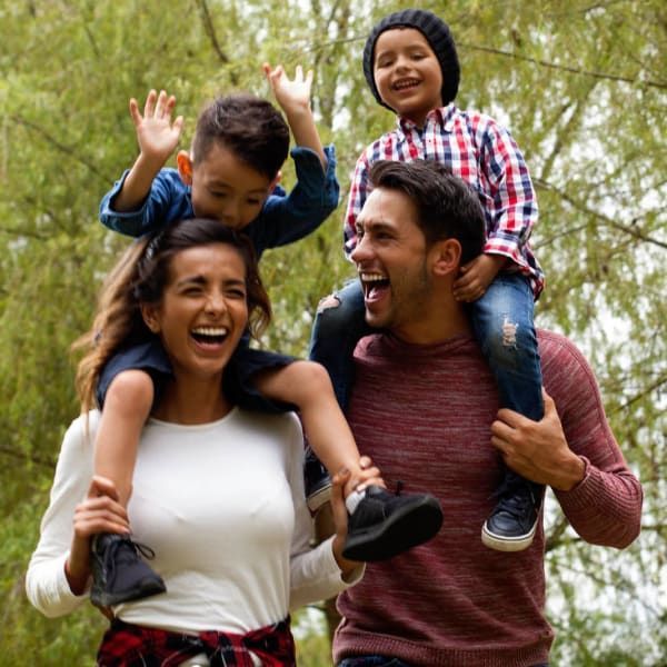 A family of four laughs outdoors as two children ride on their parents' shoulders with arms raised 