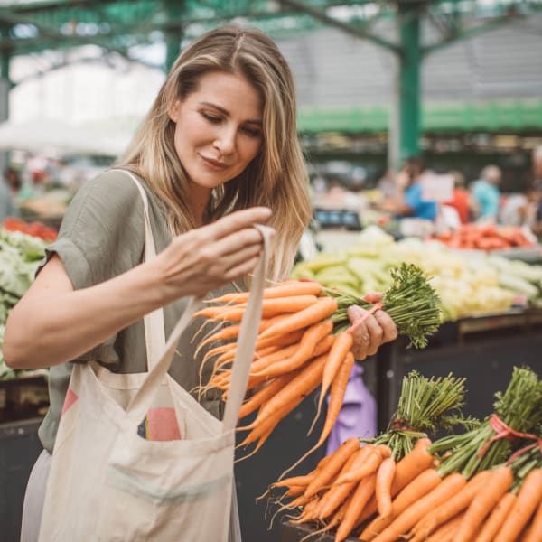 A person placing a bunch of fresh, leafy carrots into a reusable canvas tote bag at an outdoor produce market.