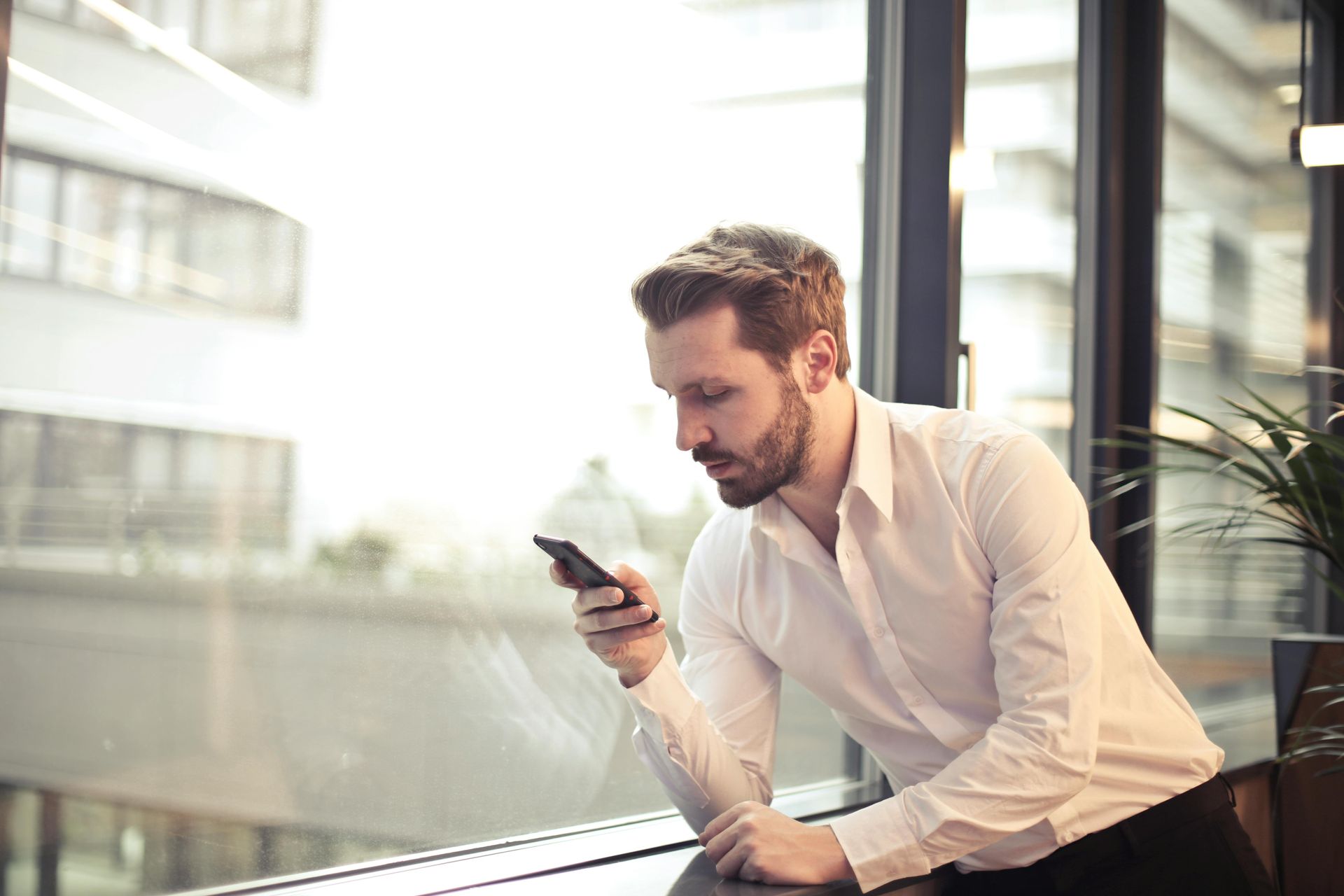 A man is sitting in front of a window looking at his cell phone.