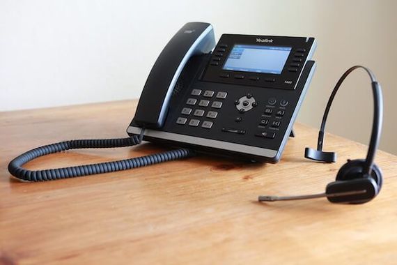 Black office phone with coiled cord, and headset resting on a wooden desk.