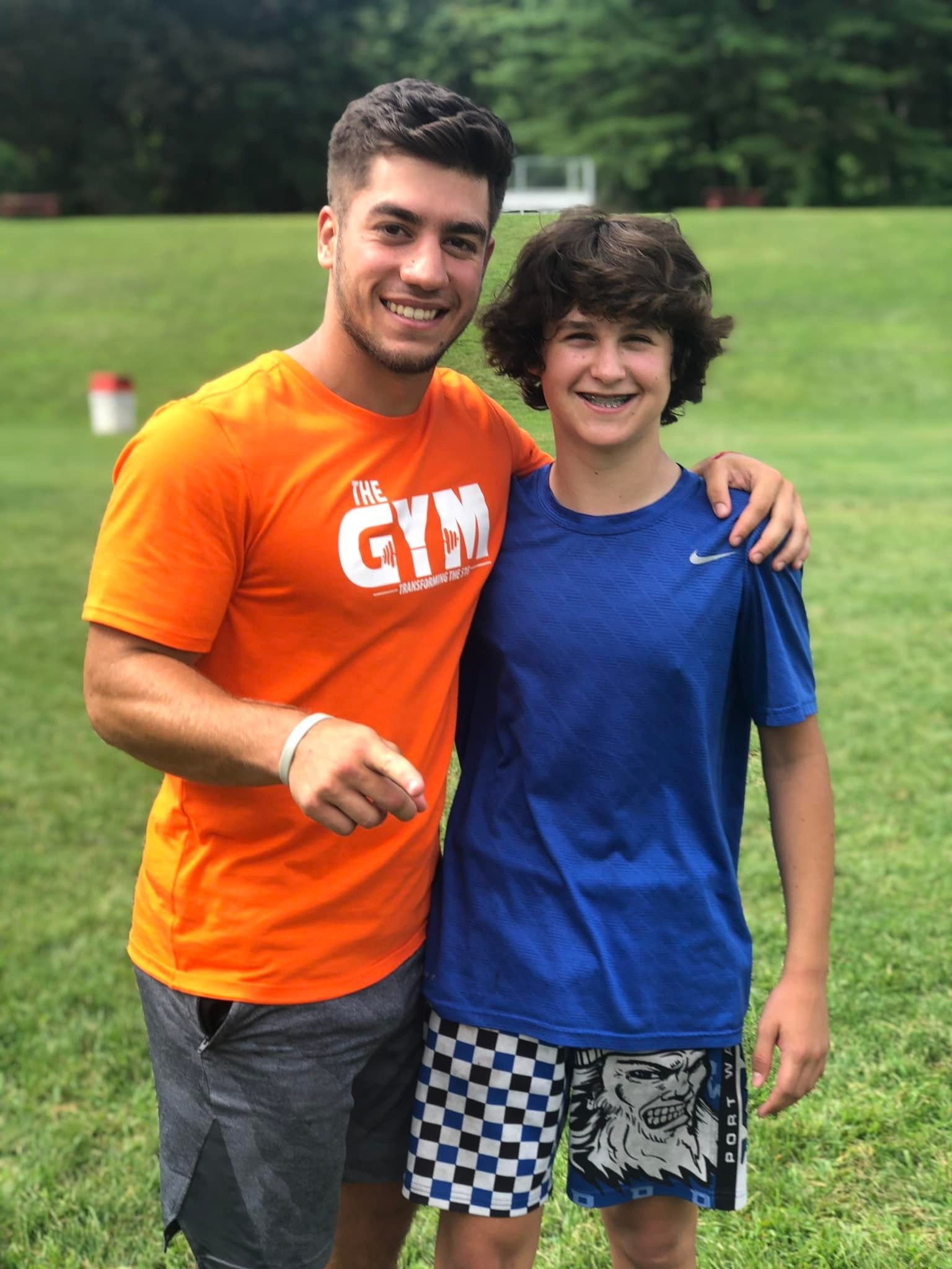 Two young men are posing for a picture in a field . one of the men is wearing an orange shirt that says gym.