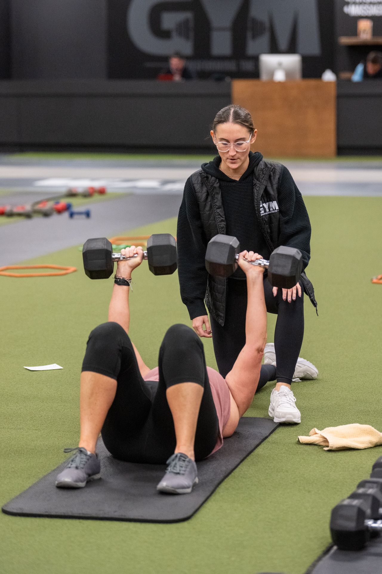 A woman is lifting dumbbells on a mat in a gym while another woman watches.