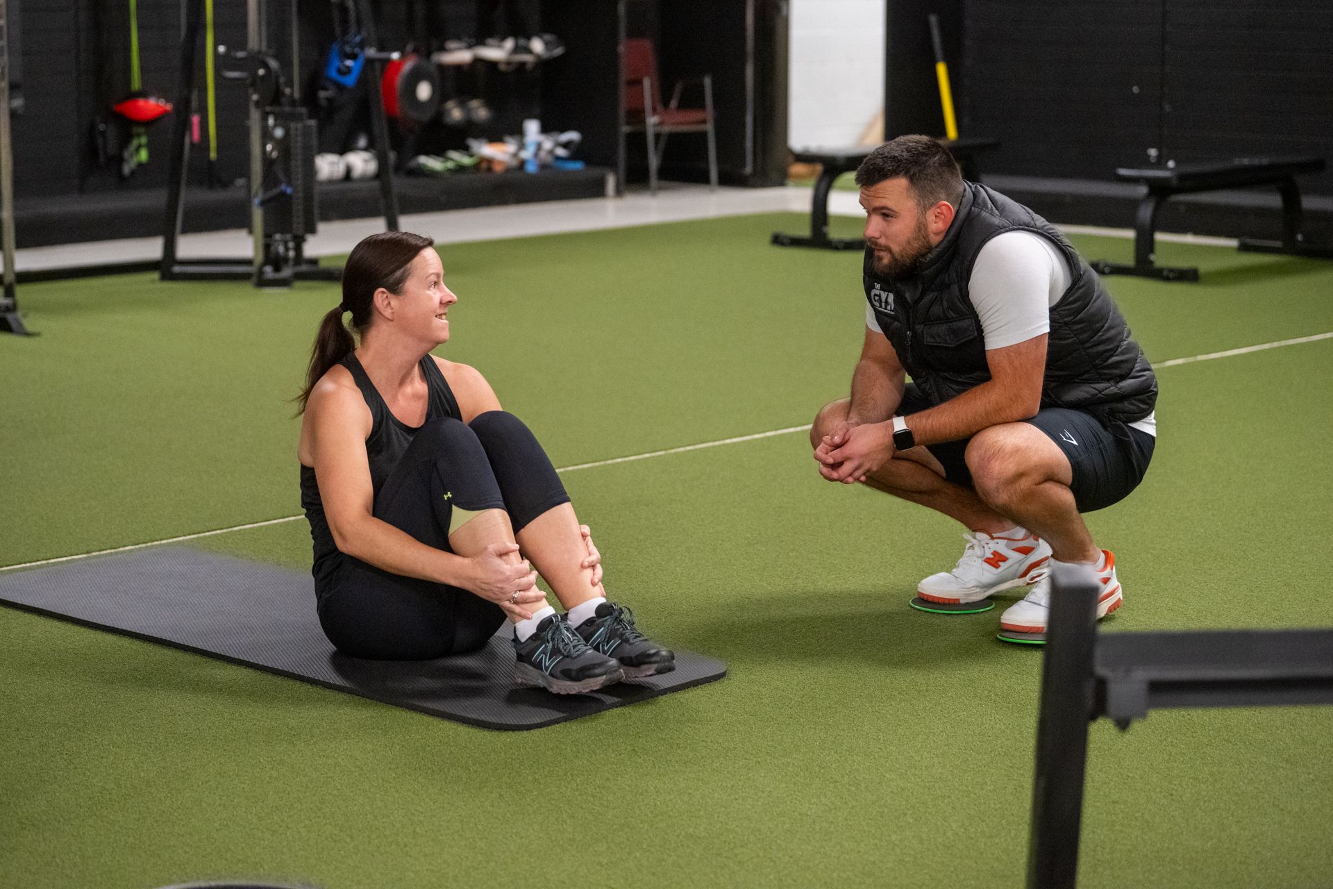 A man is squatting next to a woman sitting on a mat in a gym.