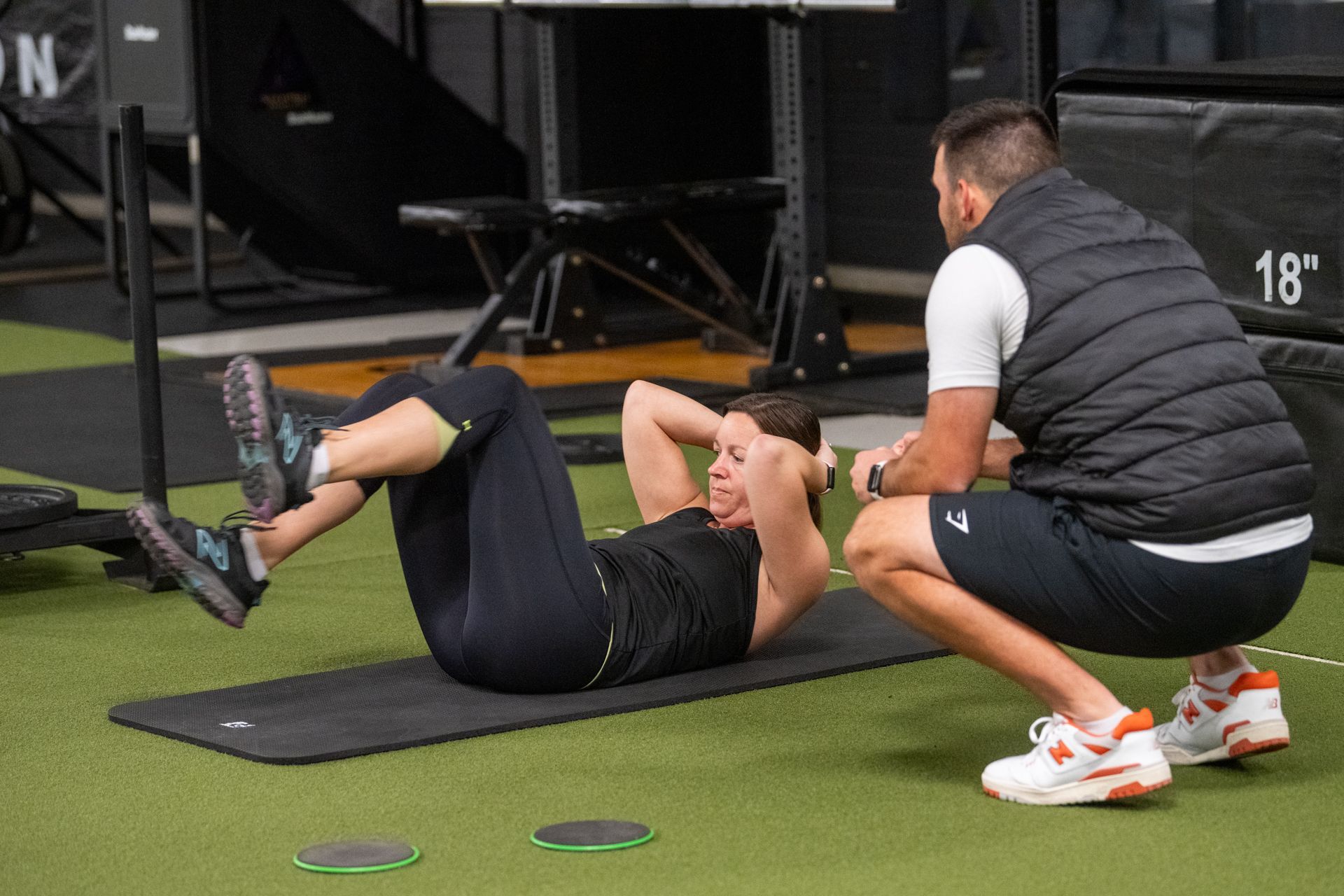 A man is squatting next to a woman doing crunches on a mat in a gym.
