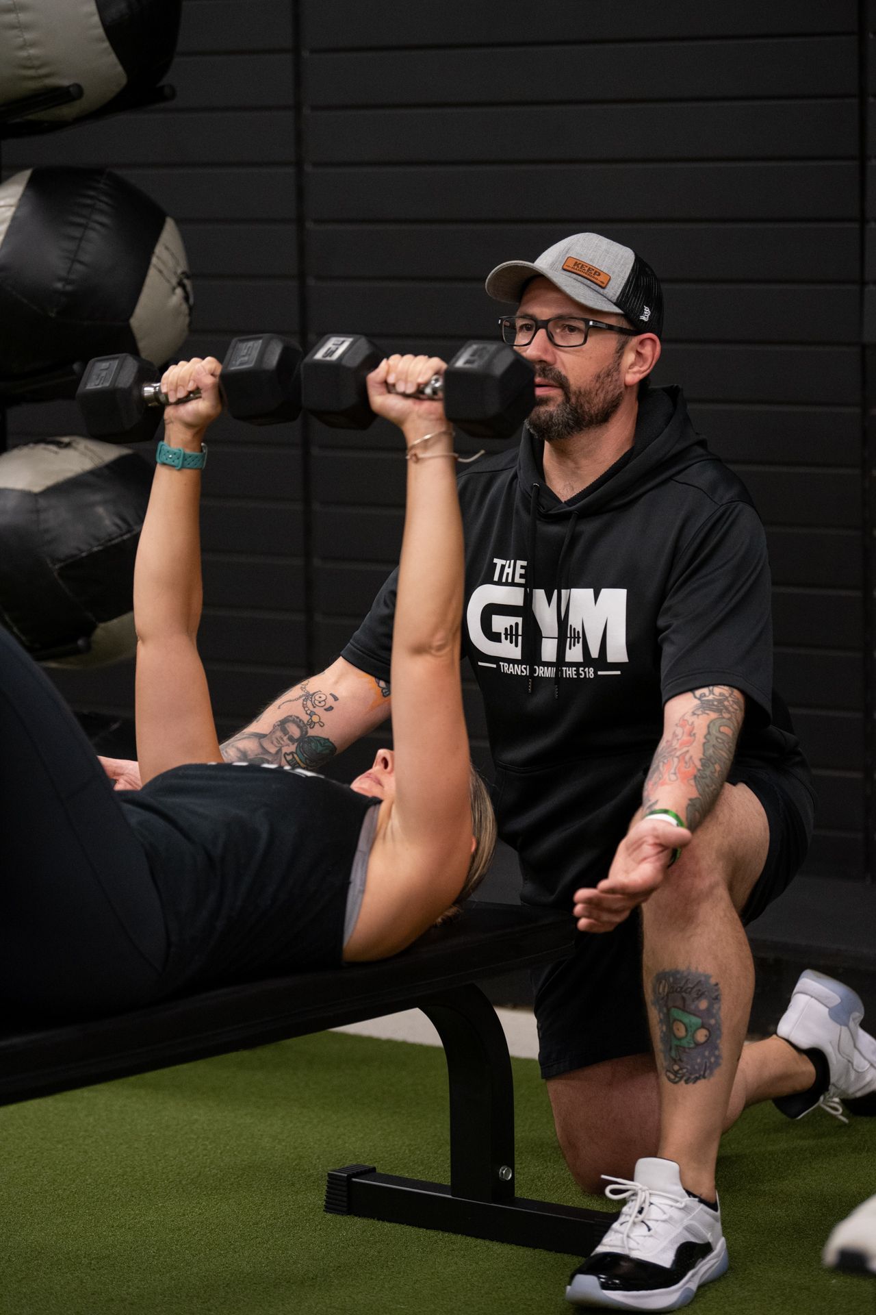 A man is helping a woman lift dumbbells on a bench in a gym.
